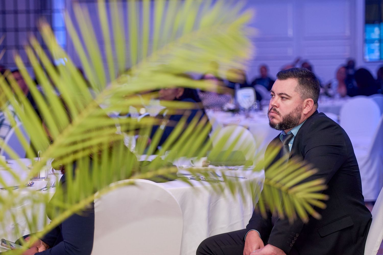 A man in a suit listens attentively at a formal event, framed by tropical palm leaves in the foreground.