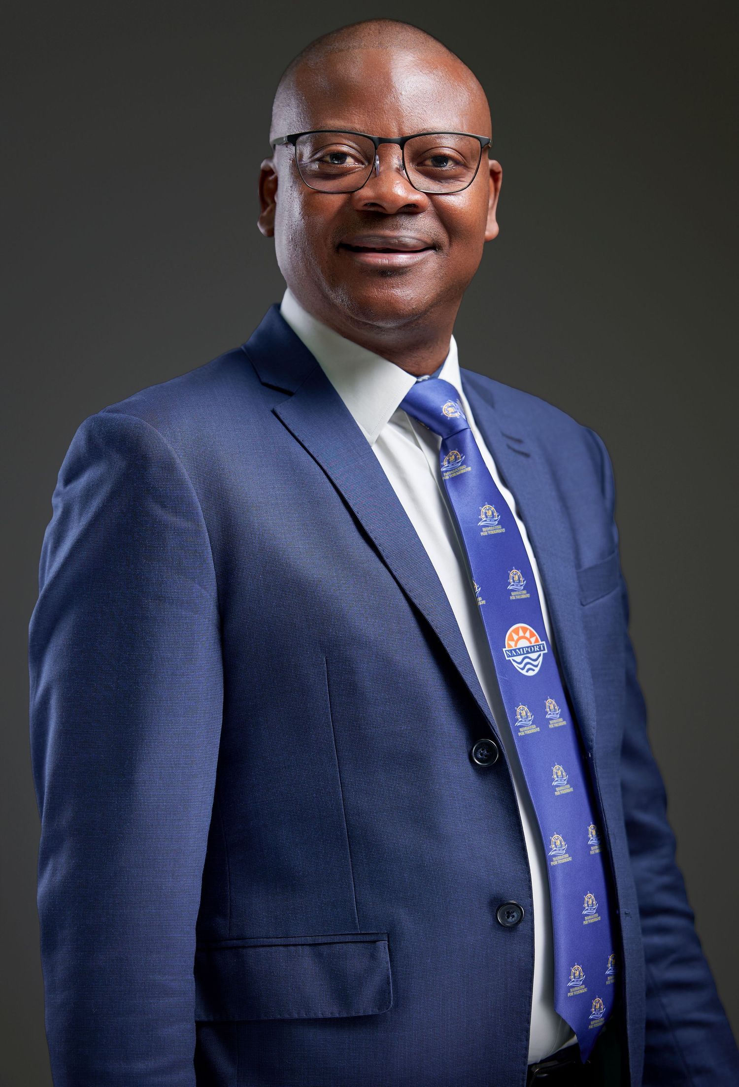 Professional headshot series showing someone in a navy blue suit and glasses against a dark background with varying poses.
