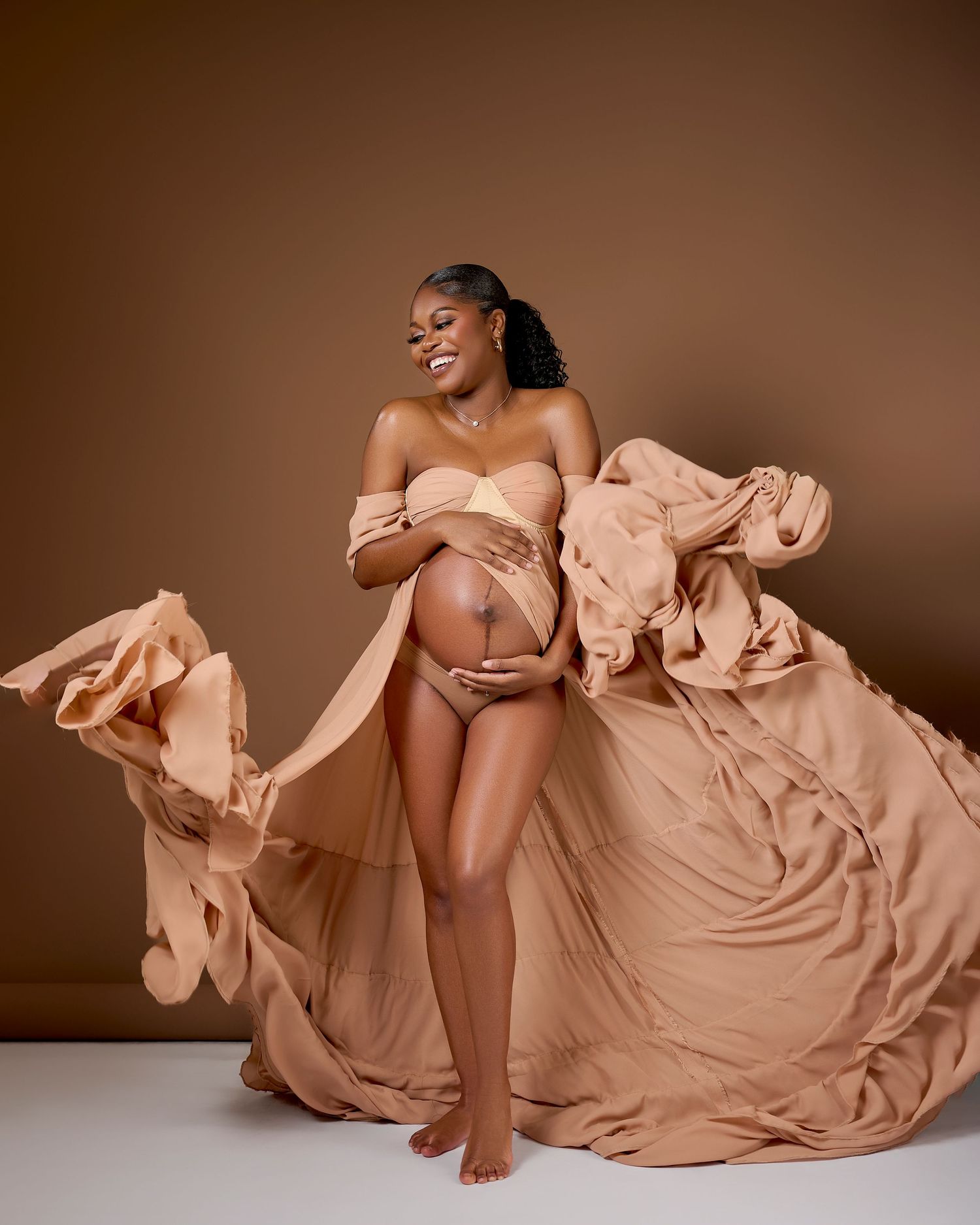 Pregnant woman in flowing peach fabric gown poses gracefully in studio maternity photoshoot against warm brown background.