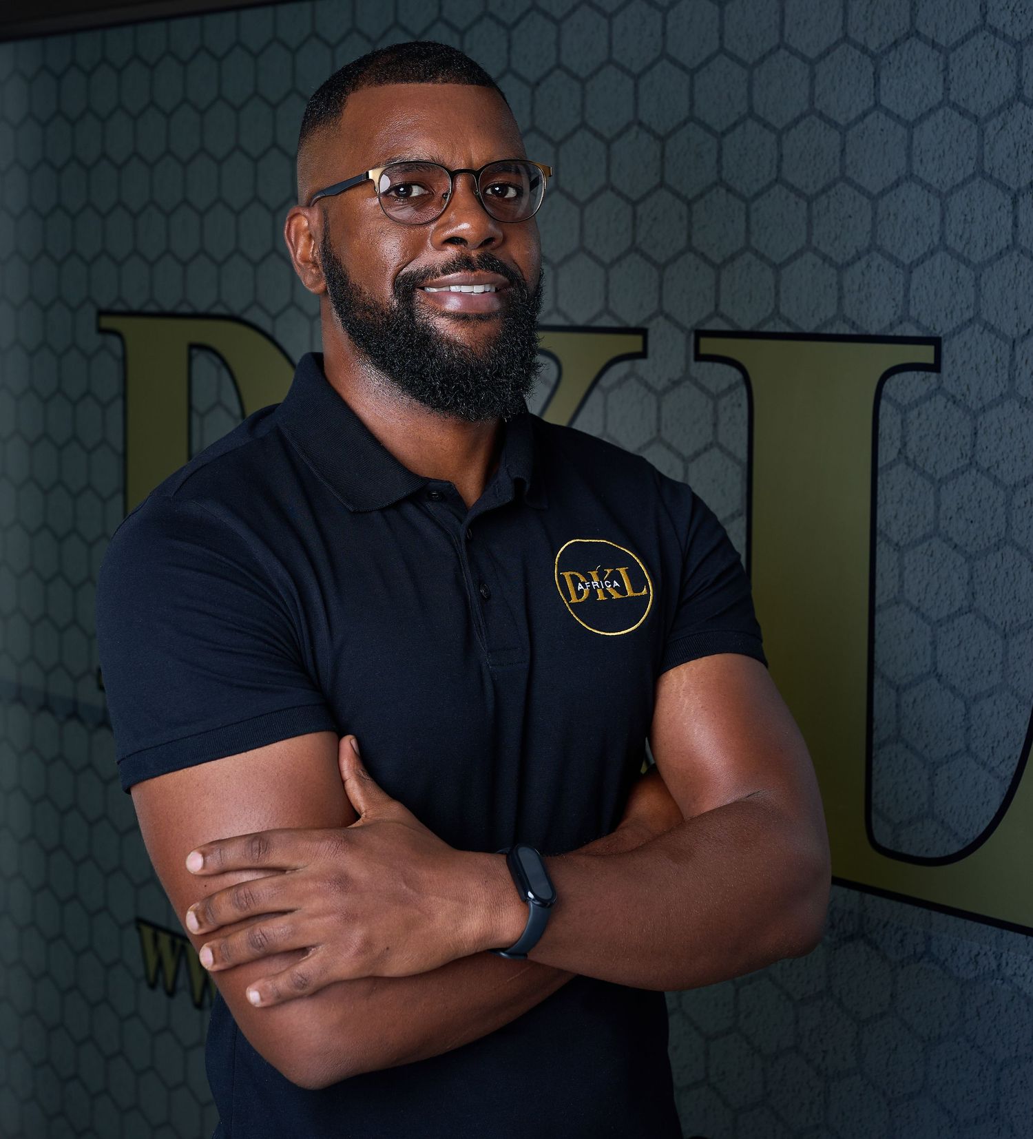 A man with a beard wearing a black polo shirt stands with his arms crossed in front of a wall with the letters 'DkL' on it.