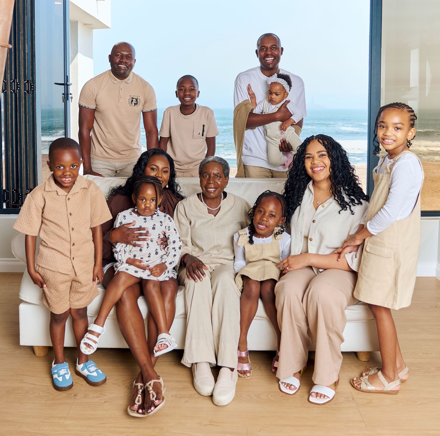 A large multigenerational Black family poses together indoors with an ocean view, wearing coordinated neutral outfits.