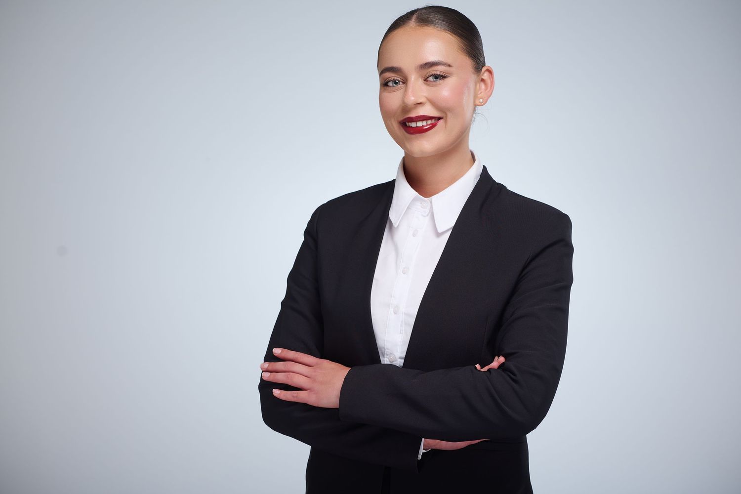 Professional businesswoman in black suit with arms crossed, smiling confidently against a light gray background.