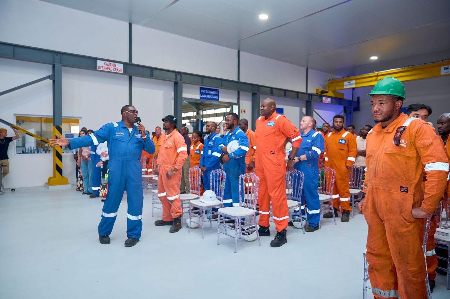 Industrial workers in orange and blue coveralls stand attentively during a facility orientation session.