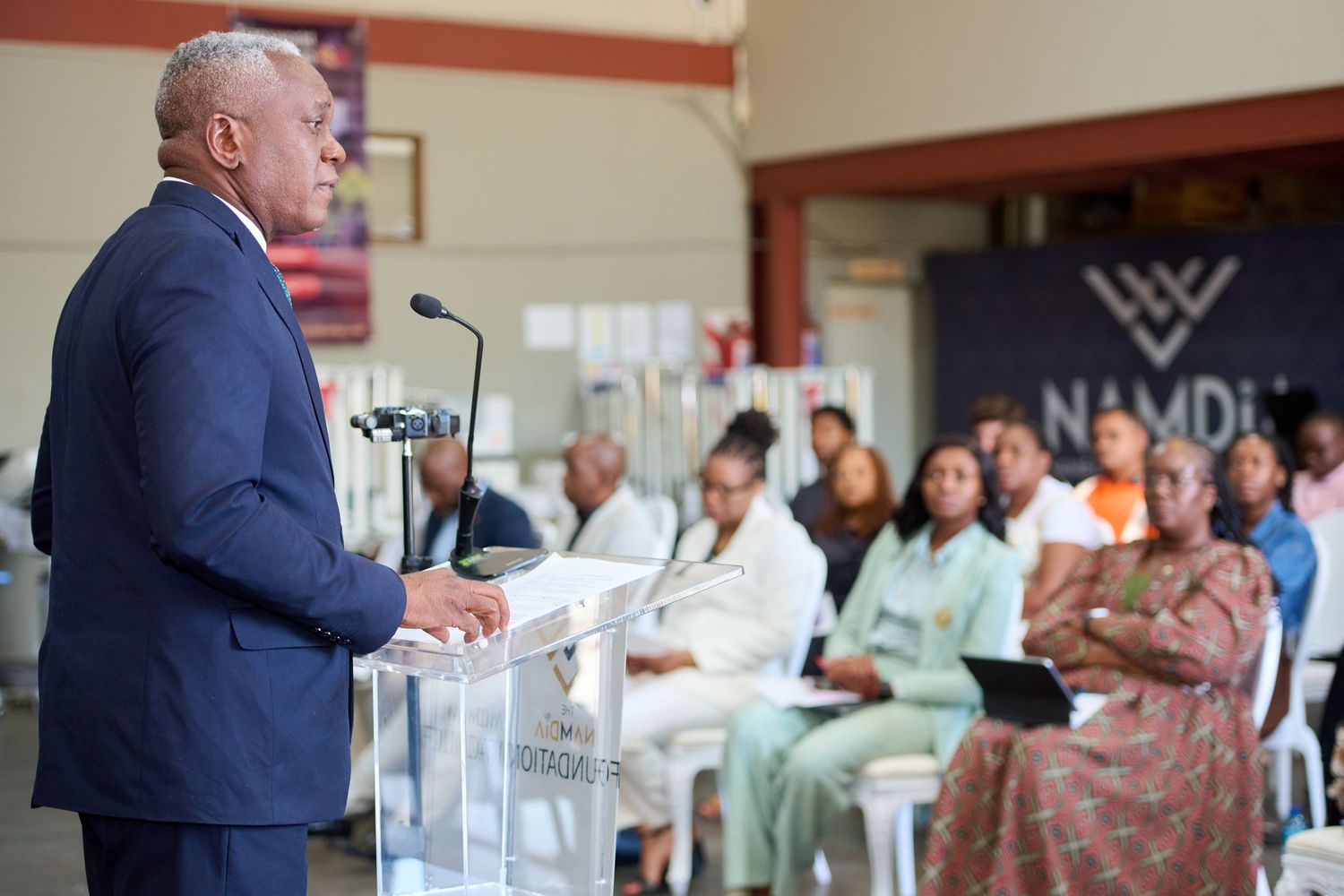 A man in a navy suit delivers a speech at a podium before a seated audience at a Namdia event.