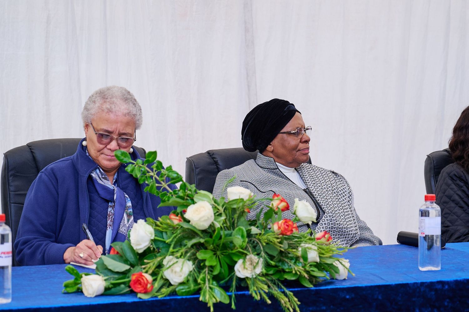Two women seated at a blue table with floral arrangement during a formal conference or meeting event.