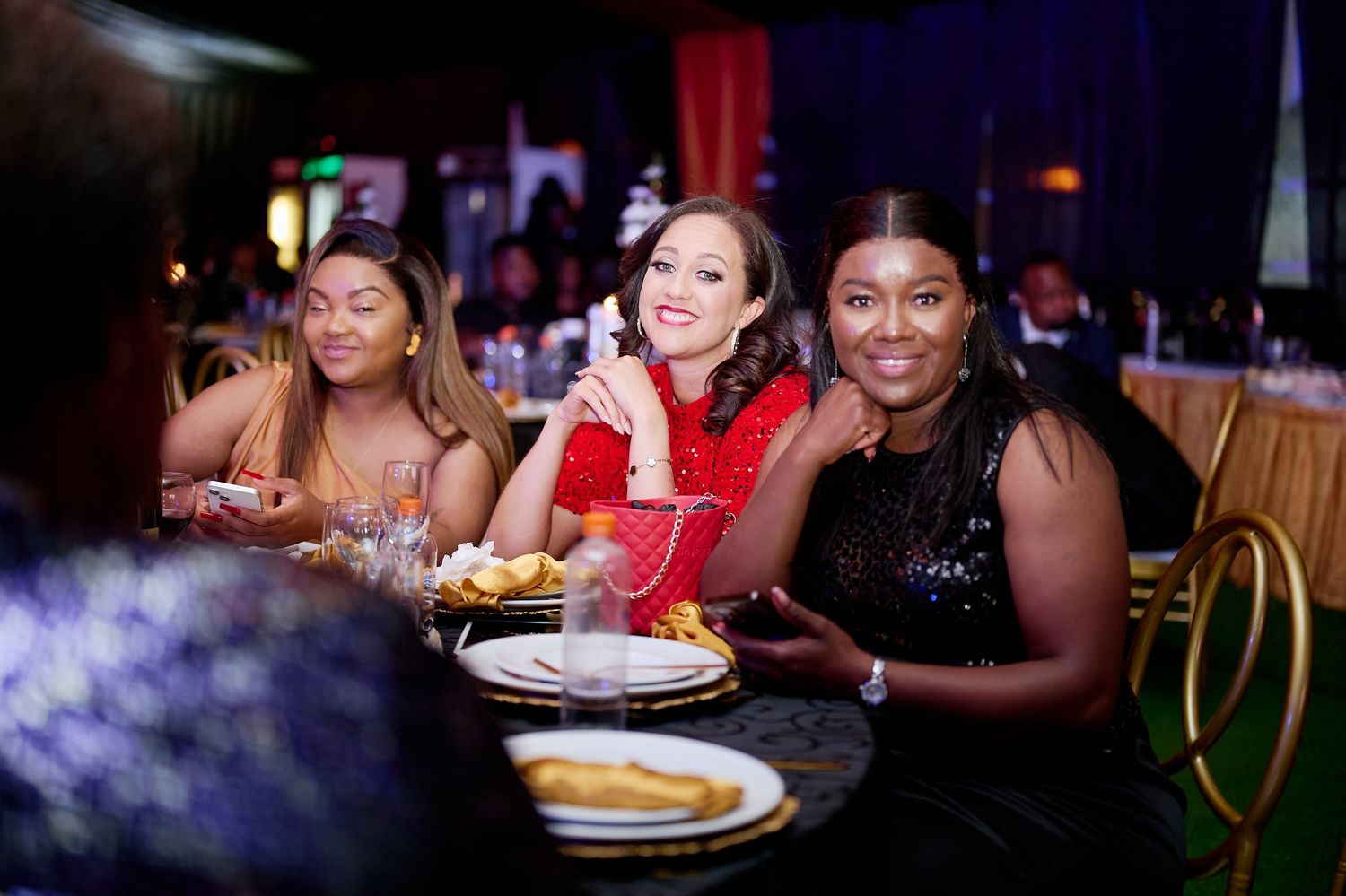 Three women smiling at a formal gala dinner table, dressed in elegant evening wear at a banquet event.