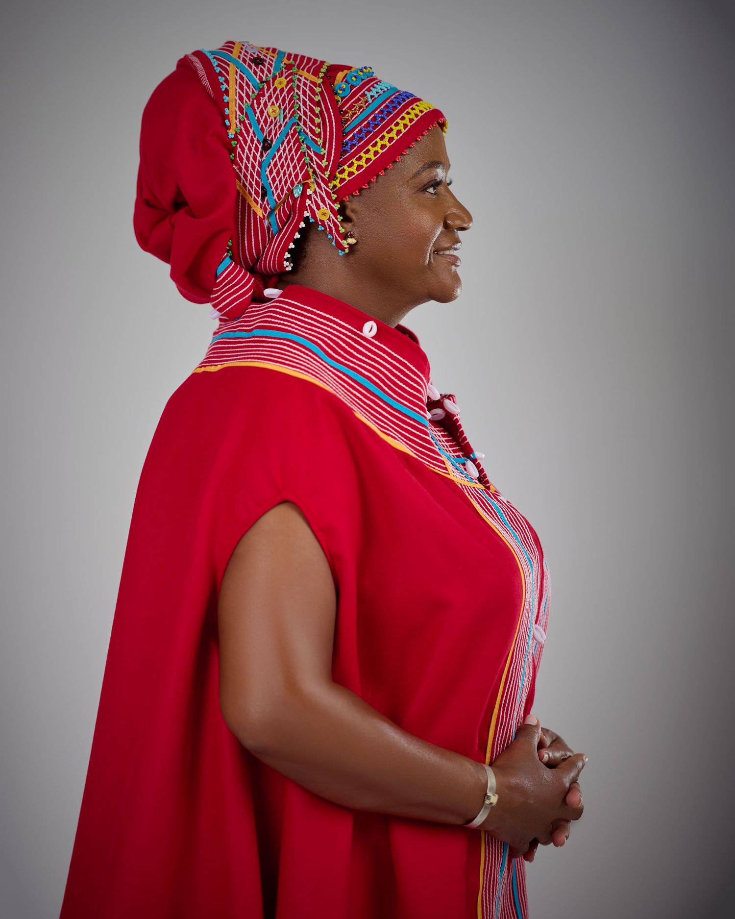 Woman in vibrant red traditional African attire with colorful beaded headwrap and necklace, smiling in profile view.