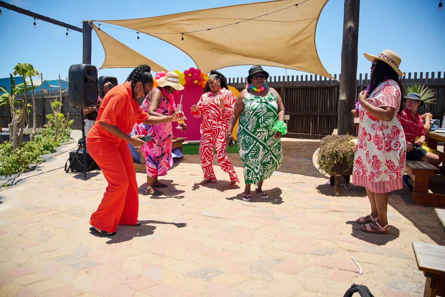 Women in vibrant tropical outfits dancing together at a sunny outdoor celebration.