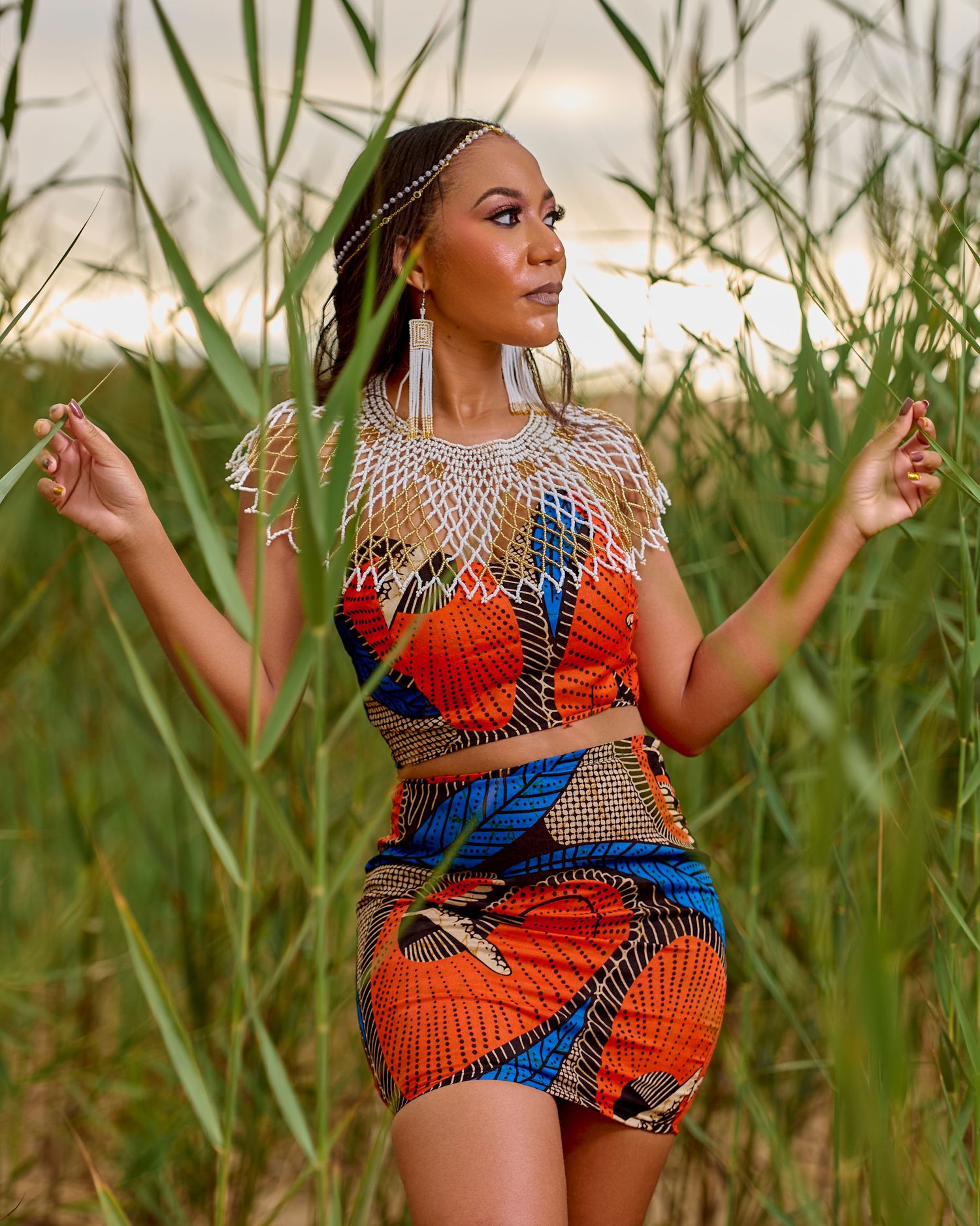 Woman in vibrant African print crop top and skirt with beaded necklace posing among tall green reeds.