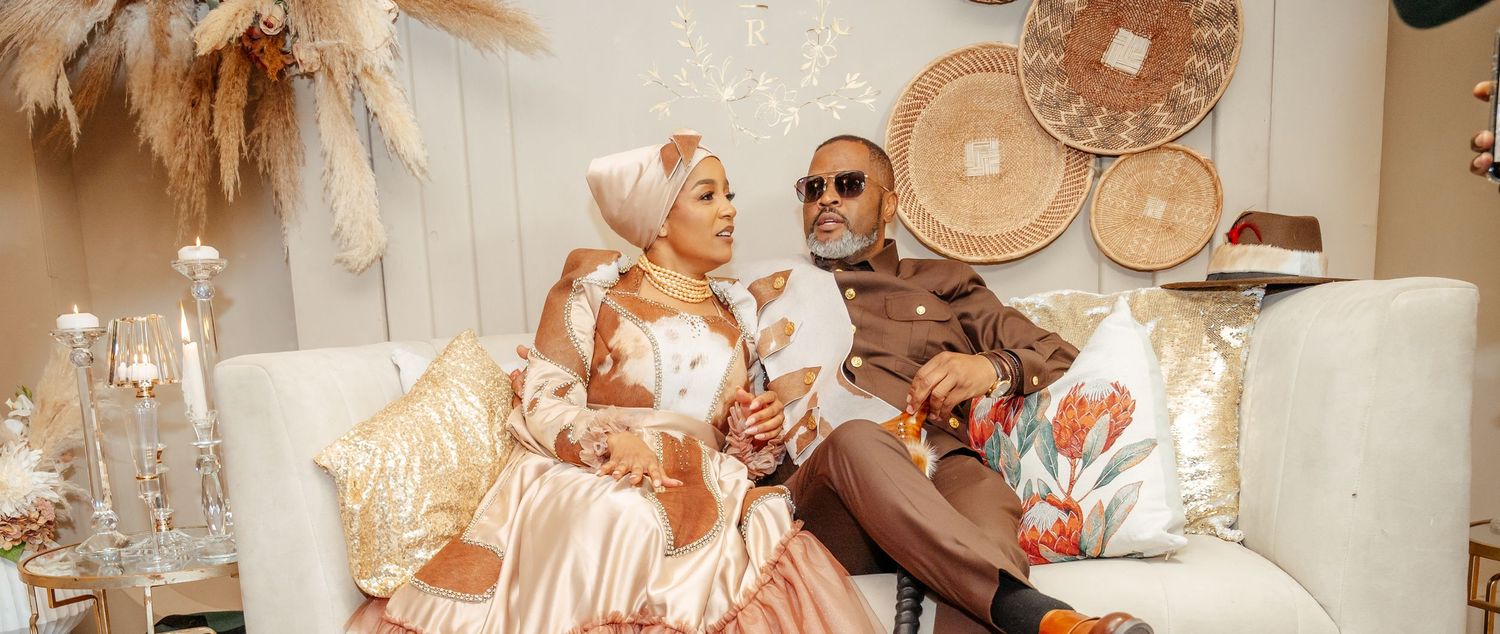 Elegant couple in traditional African attire seated on a white sofa with woven wall decor in a styled interior.