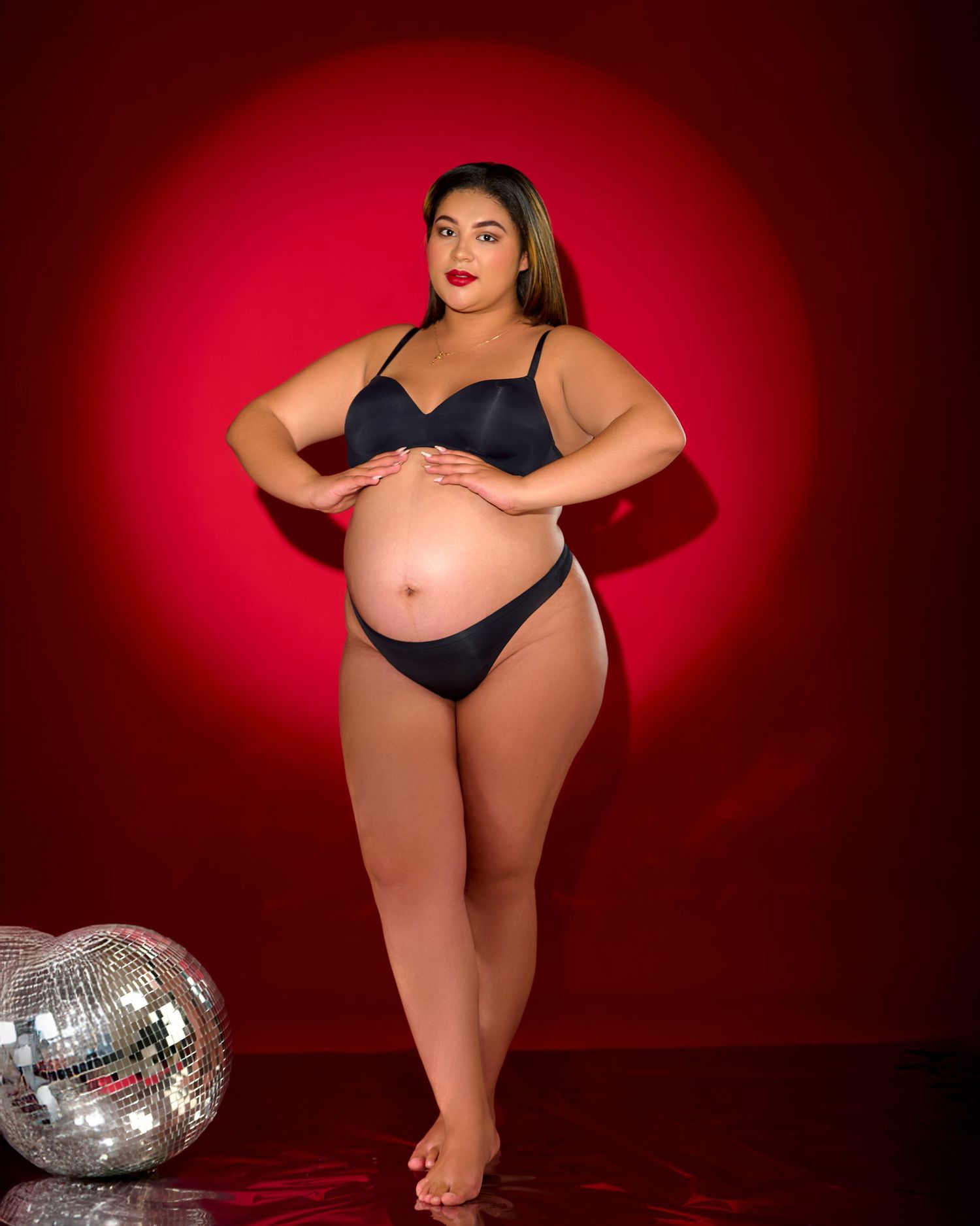 Maternity photo of a plus size model in black lingerie posing confidently against a red backdrop with a disco ball.