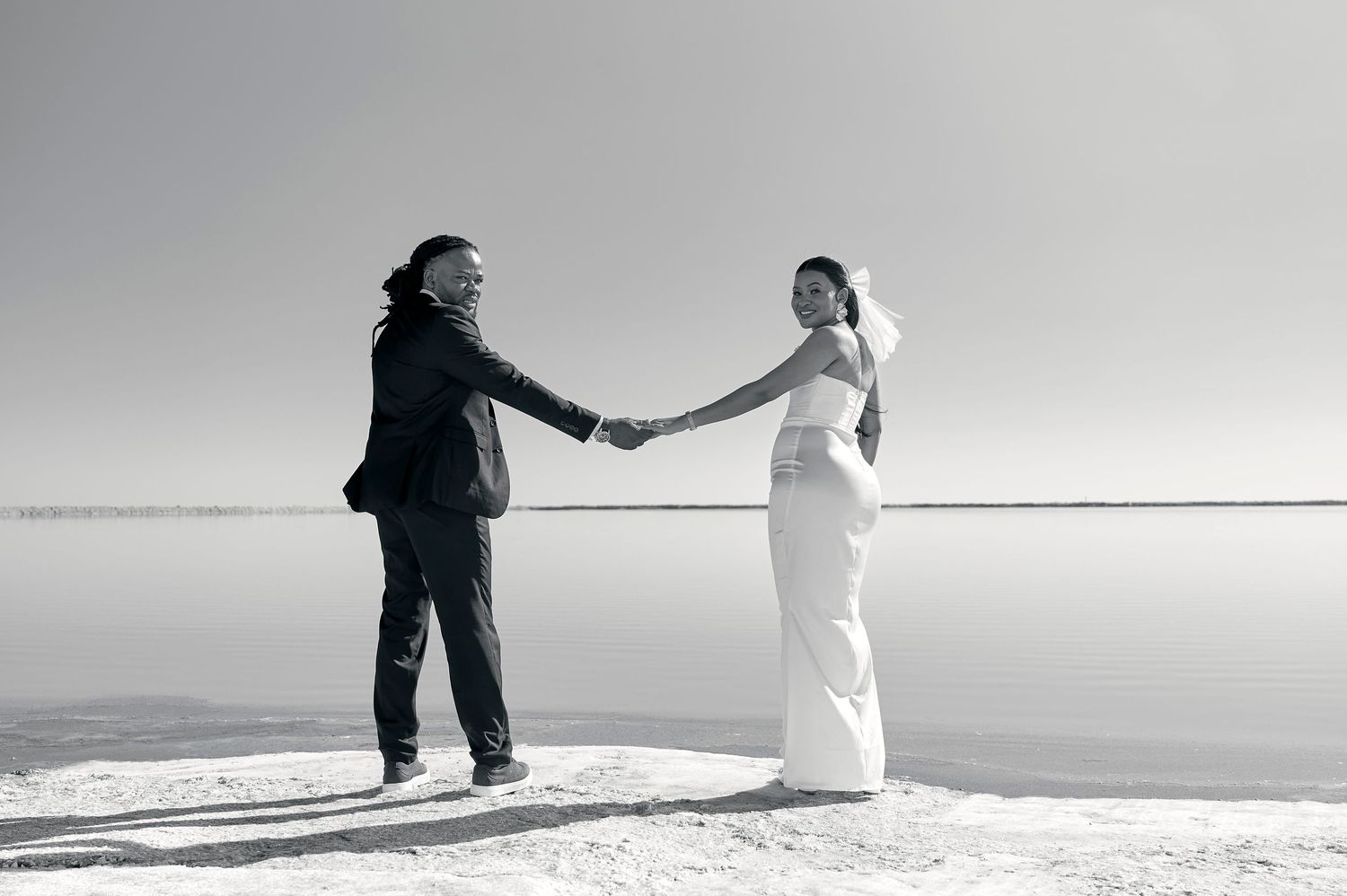 Black and white photo of a bride and groom holding hands on a salt flat, wearing wedding attire.
