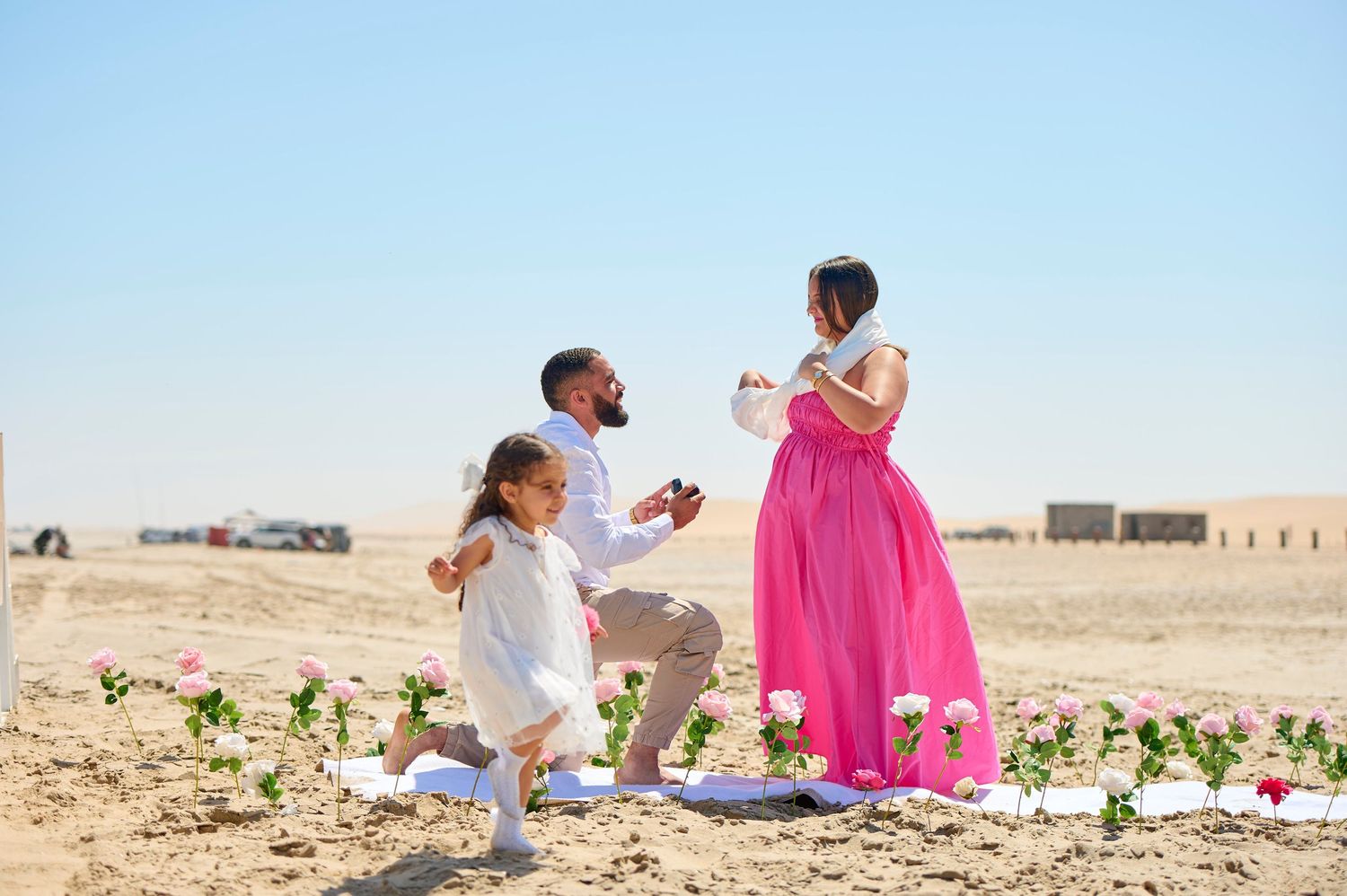 Man proposes to woman in pink dress on sandy desert with roses and young girl present.