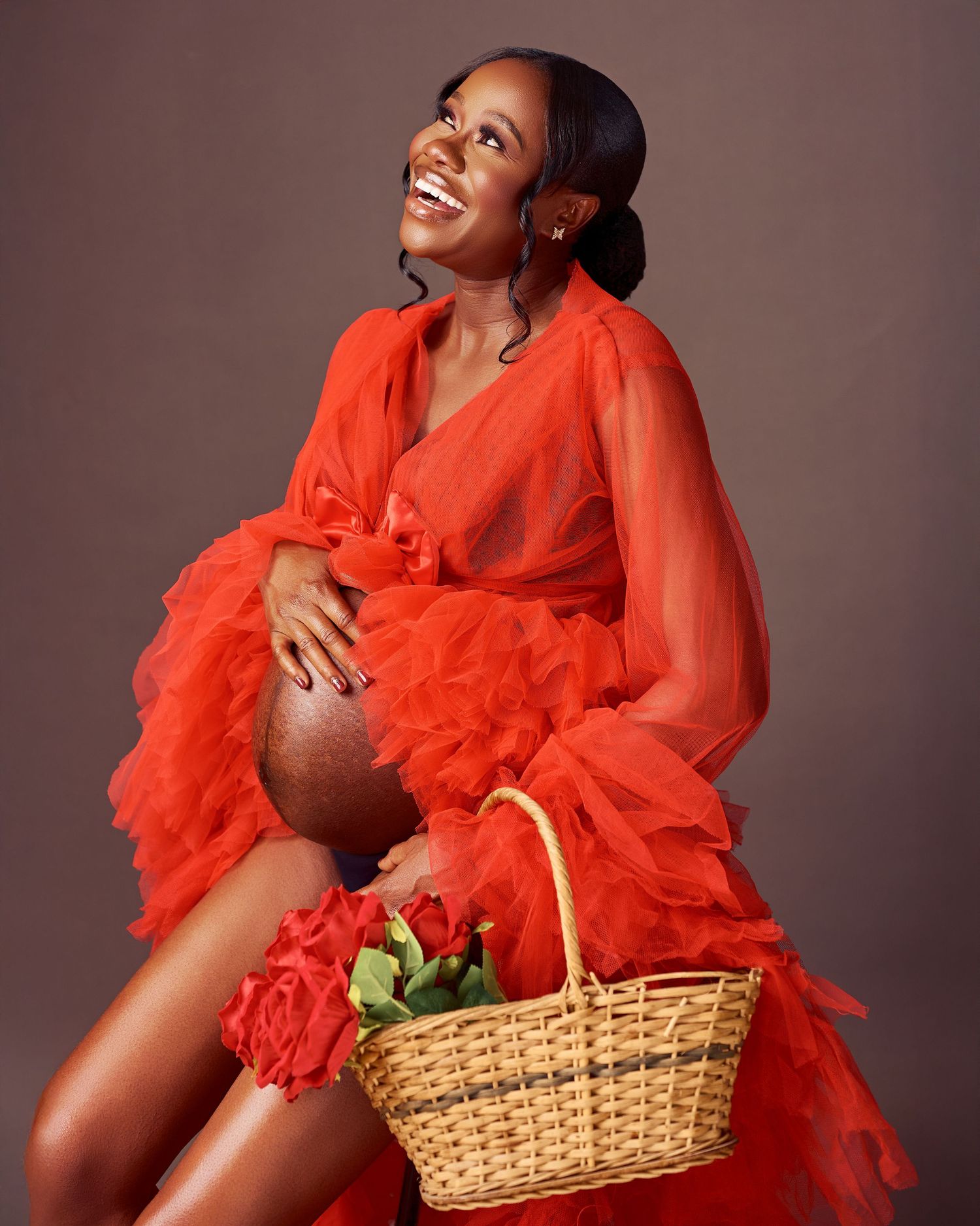A glowing expectant woman in a flowing red tulle gown poses elegantly with a wicker basket during a maternity photoshoot.