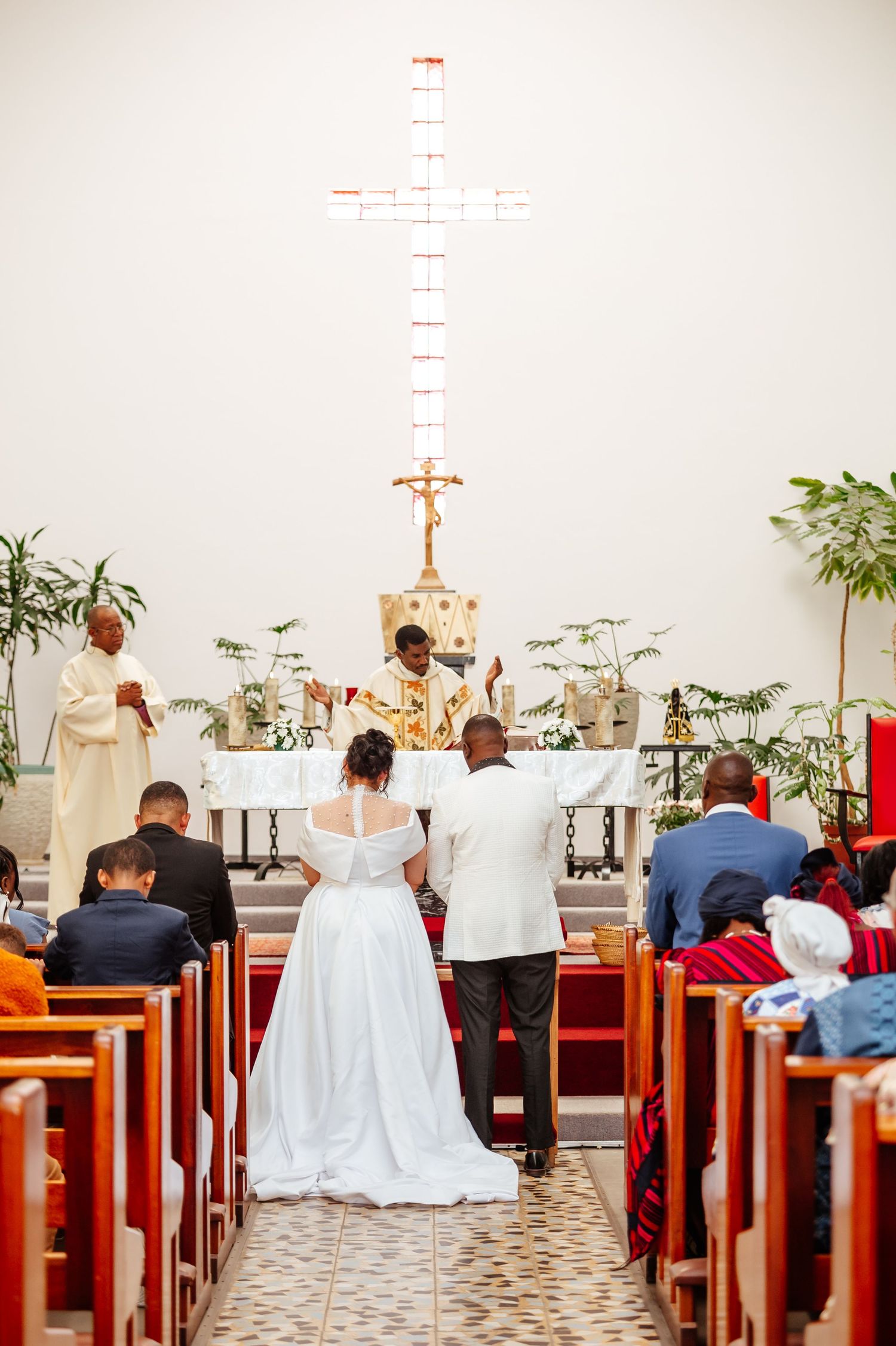 Bride and groom kneeling at church altar during wedding ceremony with priest and large cross backdrop.