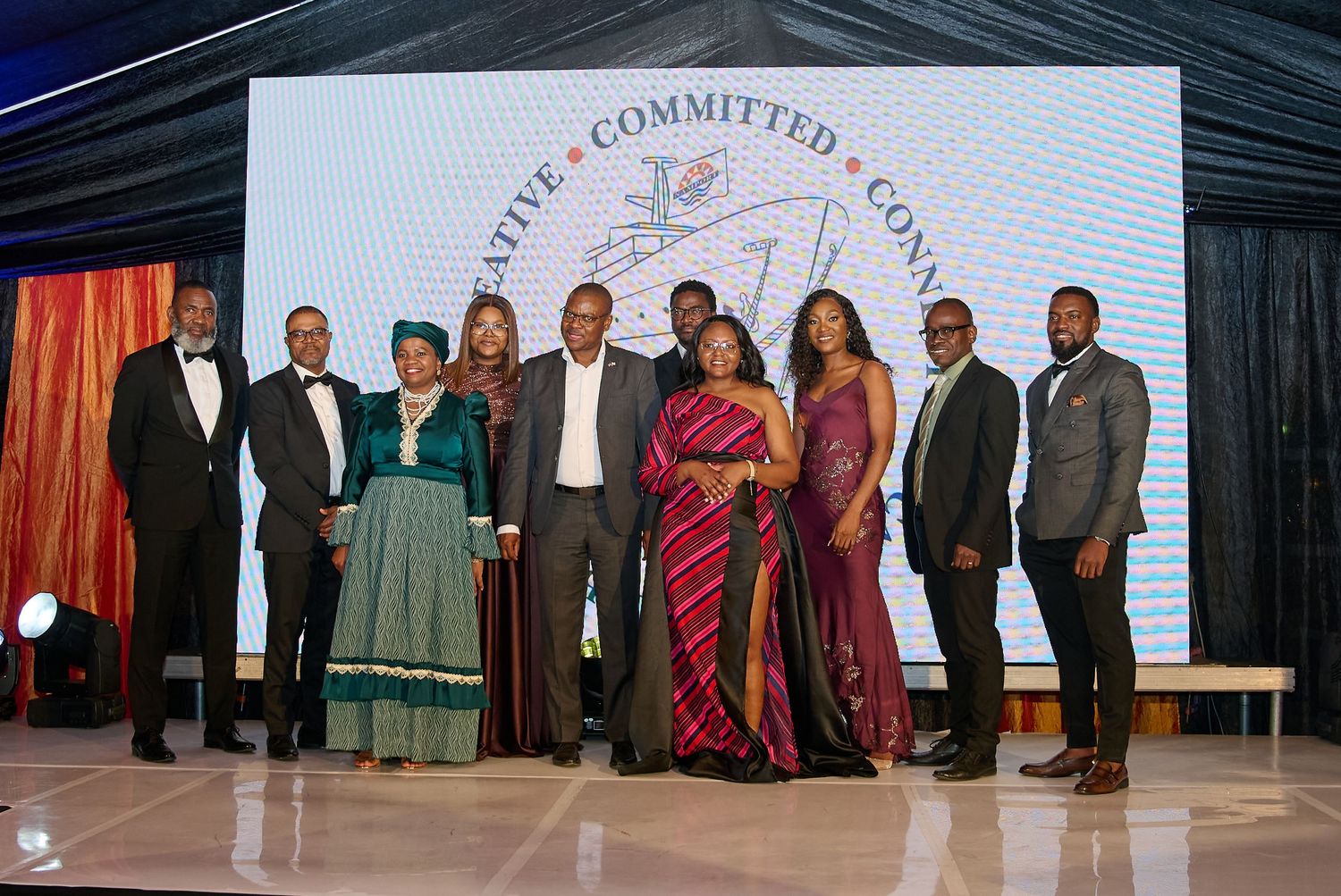 Group of elegantly dressed professionals posing together at a formal gala event in front of an illuminated backdrop.