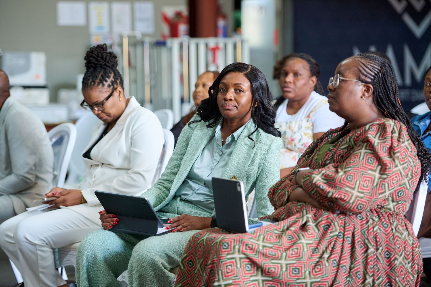 Three women sitting in chairs at a conference, two holding tablets, listening attentively to a presentation.