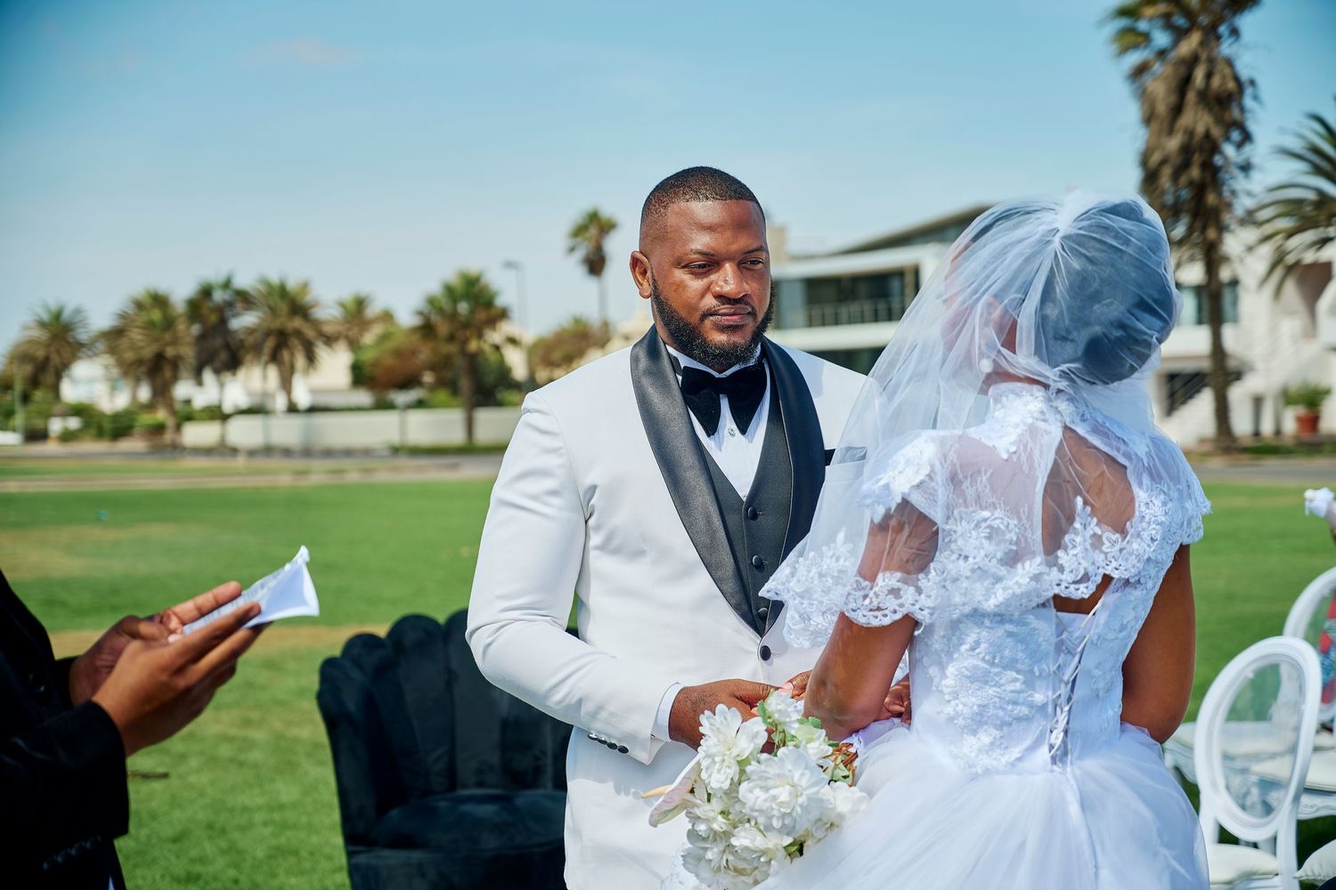 A groom in a white tuxedo exchanges vows with his bride in a white lace gown outdoors on a green lawn.