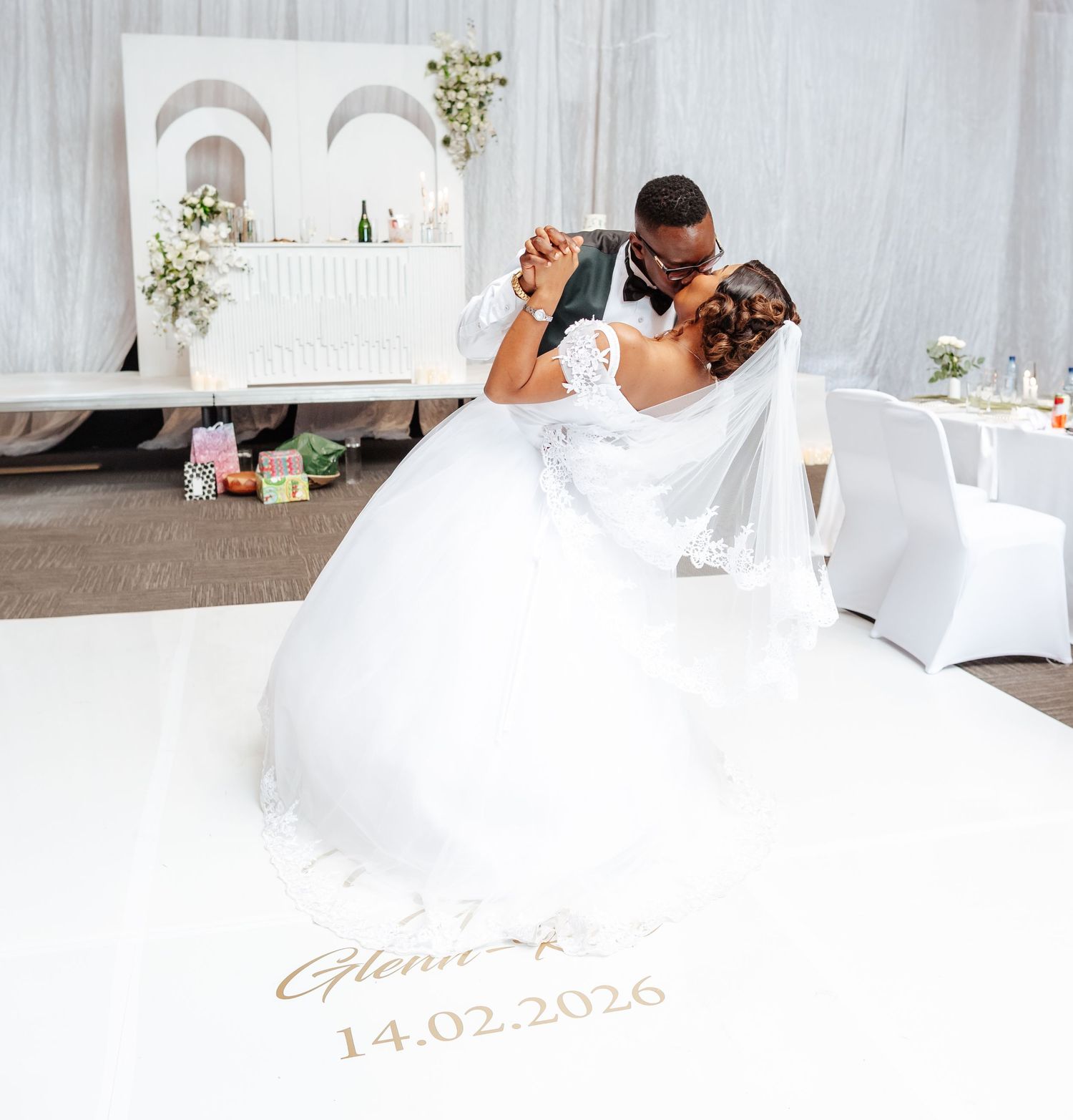 Couple sharing first dance kiss on personalized white dance floor reading 14.02.2026 at elegant wedding reception.