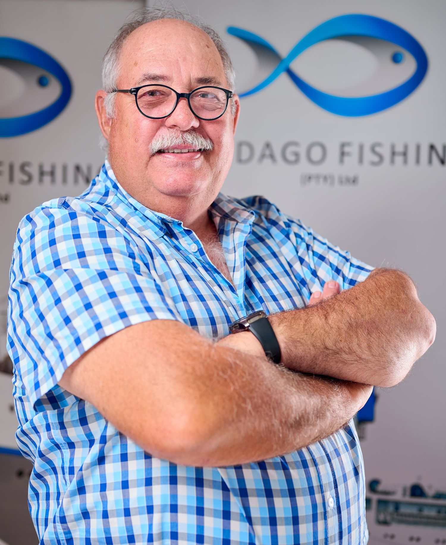 Person in blue checkered shirt and glasses posing with crossed arms against white wall with logo.