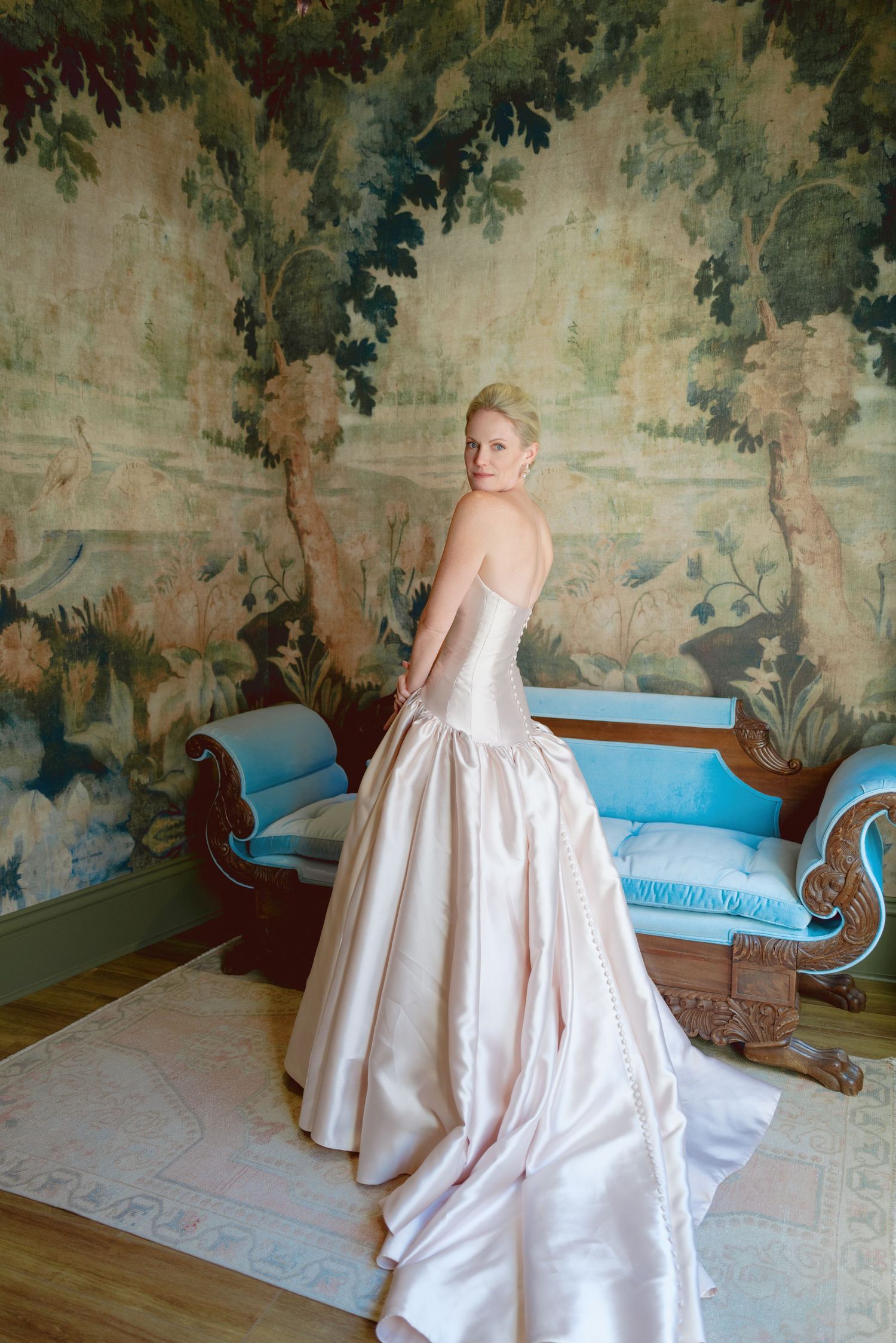 Blonde bride in strapless blush satin ballgown poses elegantly before ornate tapestry wall beside blue antique settee.