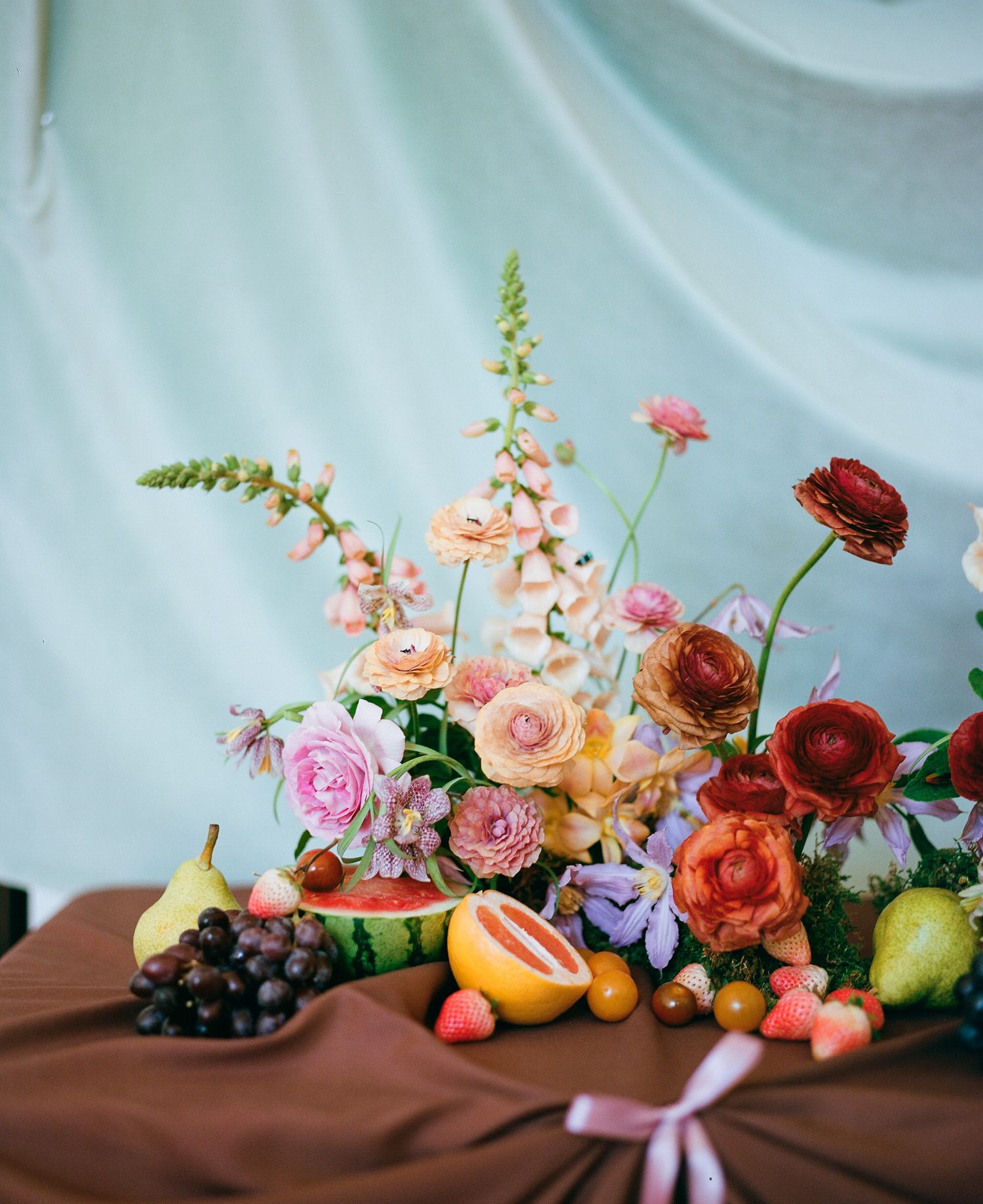 A floral arrangement with roses, grapes, and citrus fruits against a blue and brown backdrop creates an elegant still life.