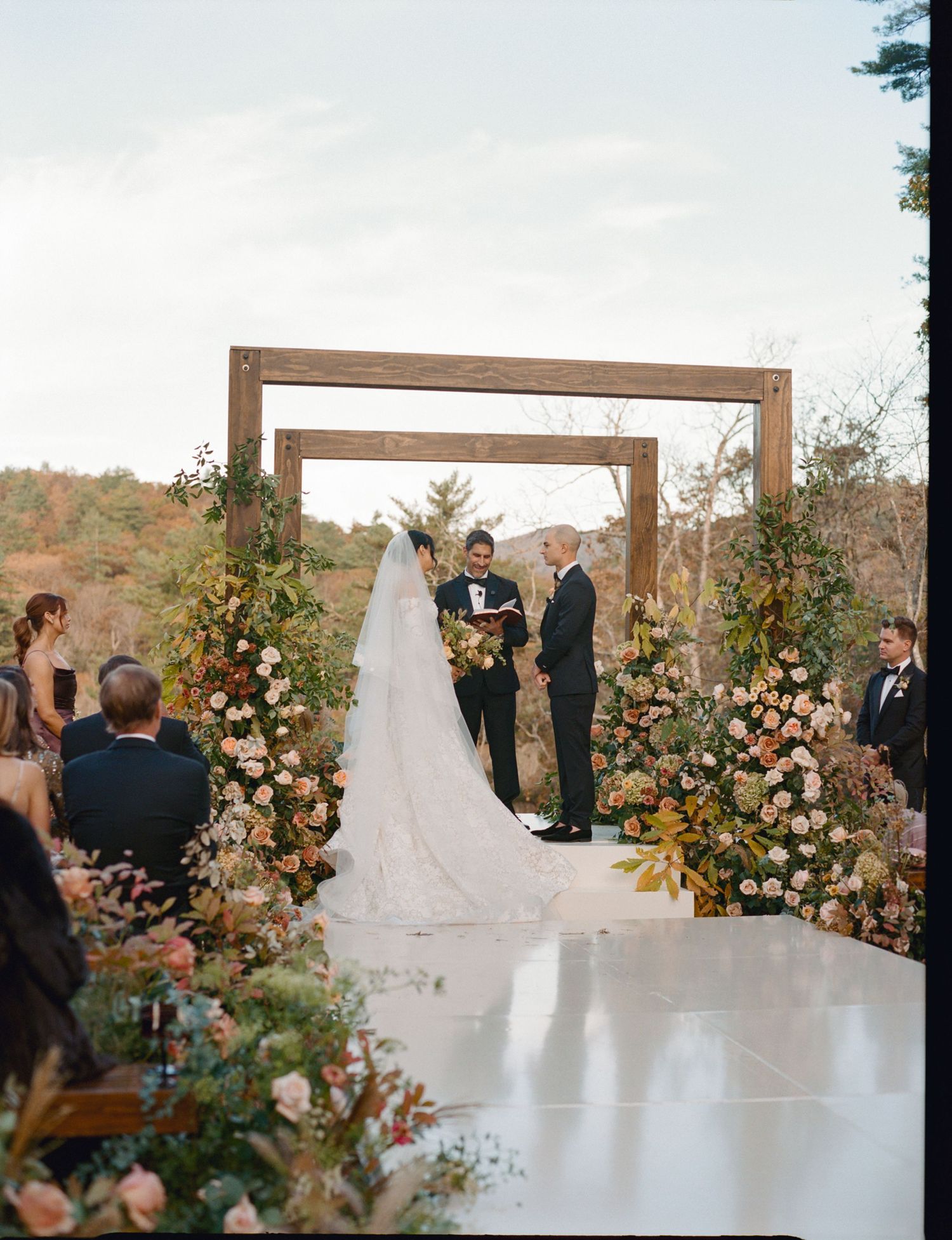Outdoor wedding ceremony with floral arch, bride and groom at altar, lush pink blooms, wooden frame, scenic nature backdrop.