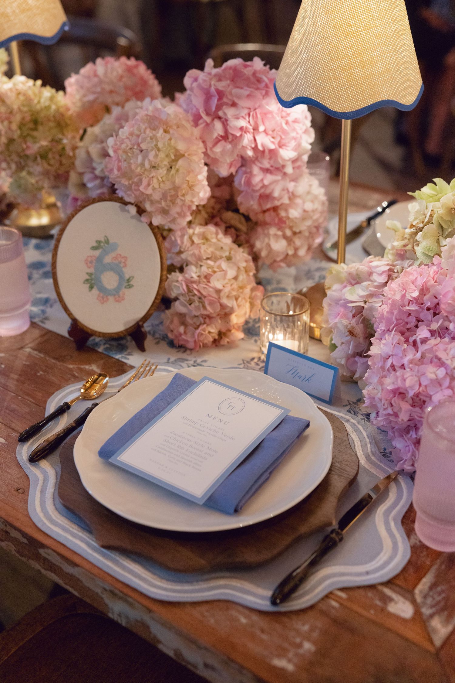 Elegant table settings with pink hydrangea centerpieces and blue accents on rustic wooden tables at a formal event.