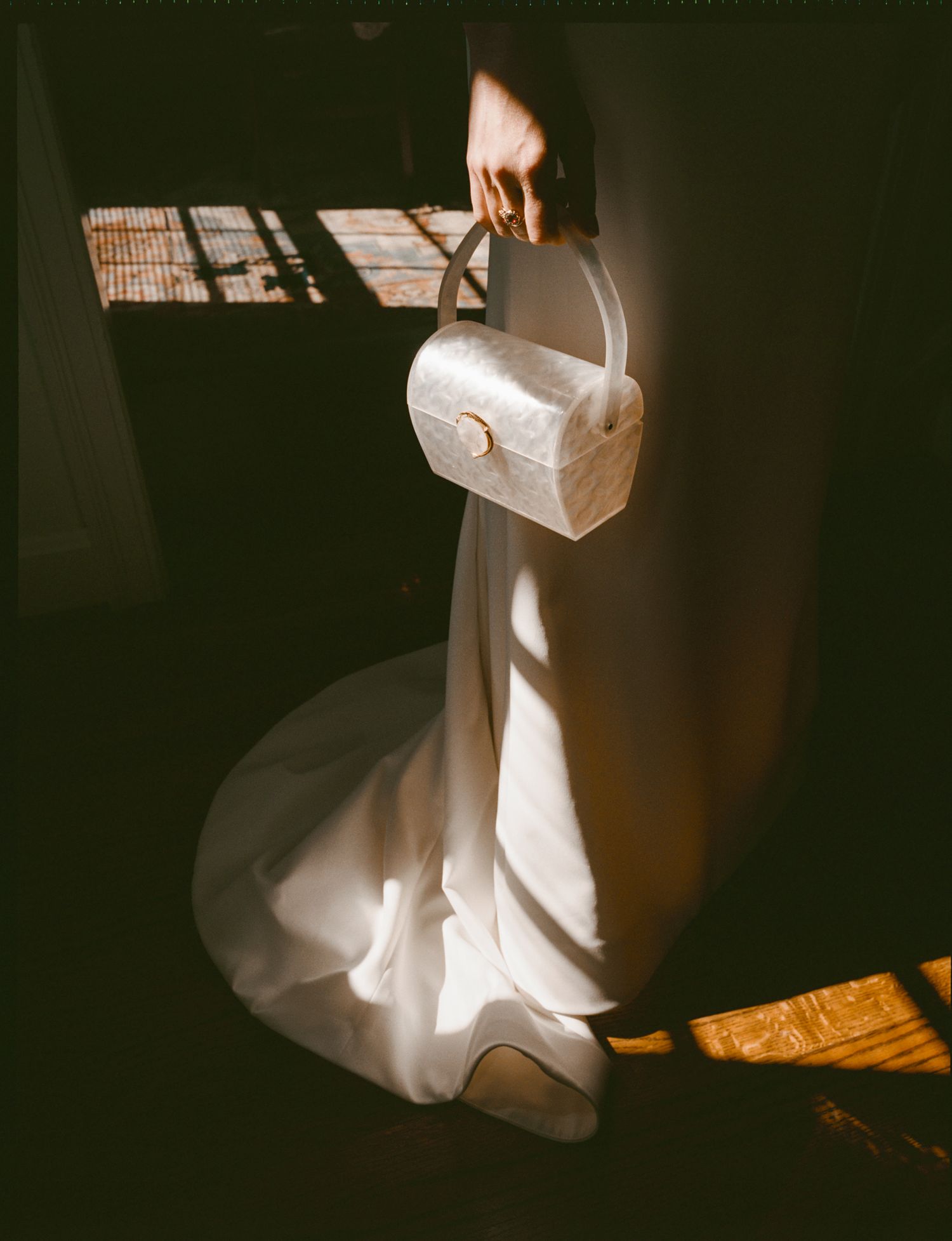 Bride holding a white structured handbag with matching heels, illuminated by dramatic sunlight against a dark background.