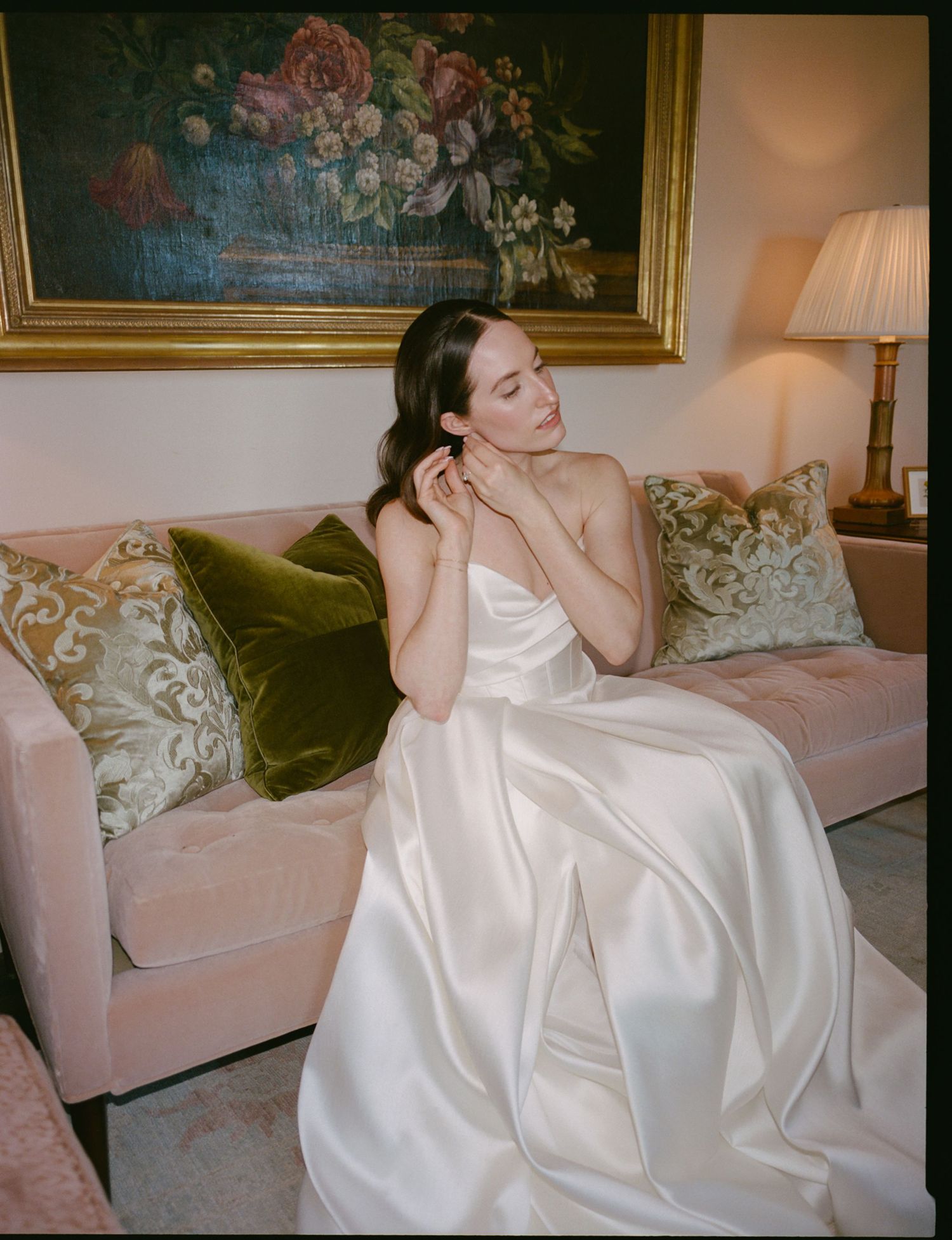 Bride in ivory satin strapless ball gown seated on pink sofa with green velvet pillows beneath floral painting.