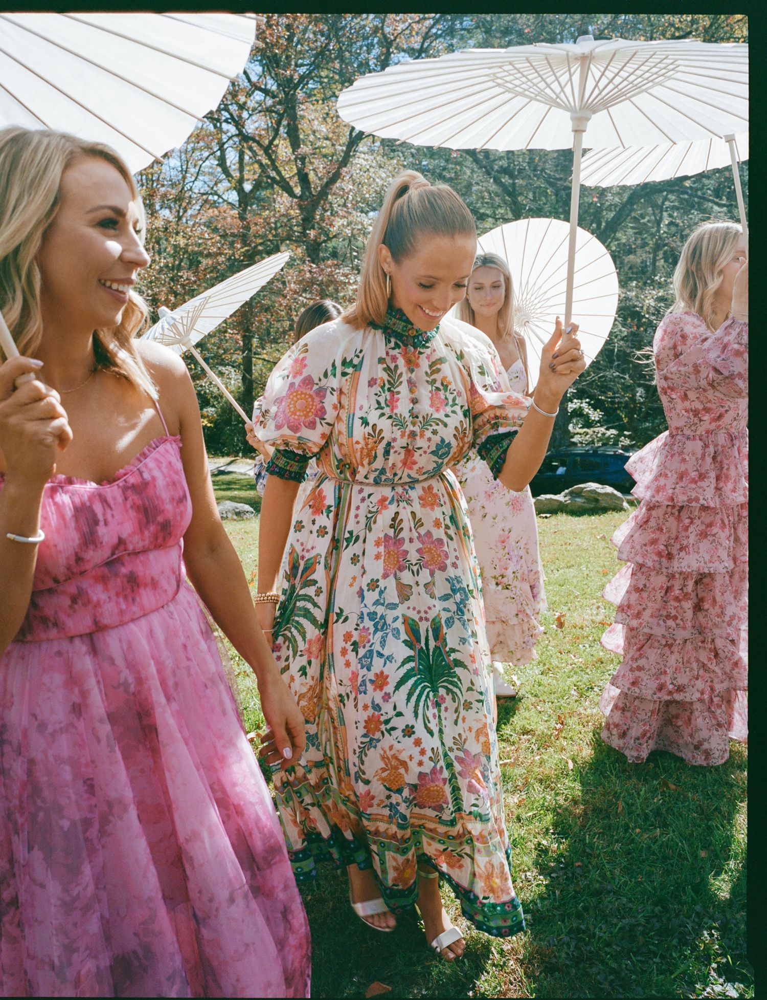 Women in floral and pink dresses holding white parasols at an outdoor garden party event.