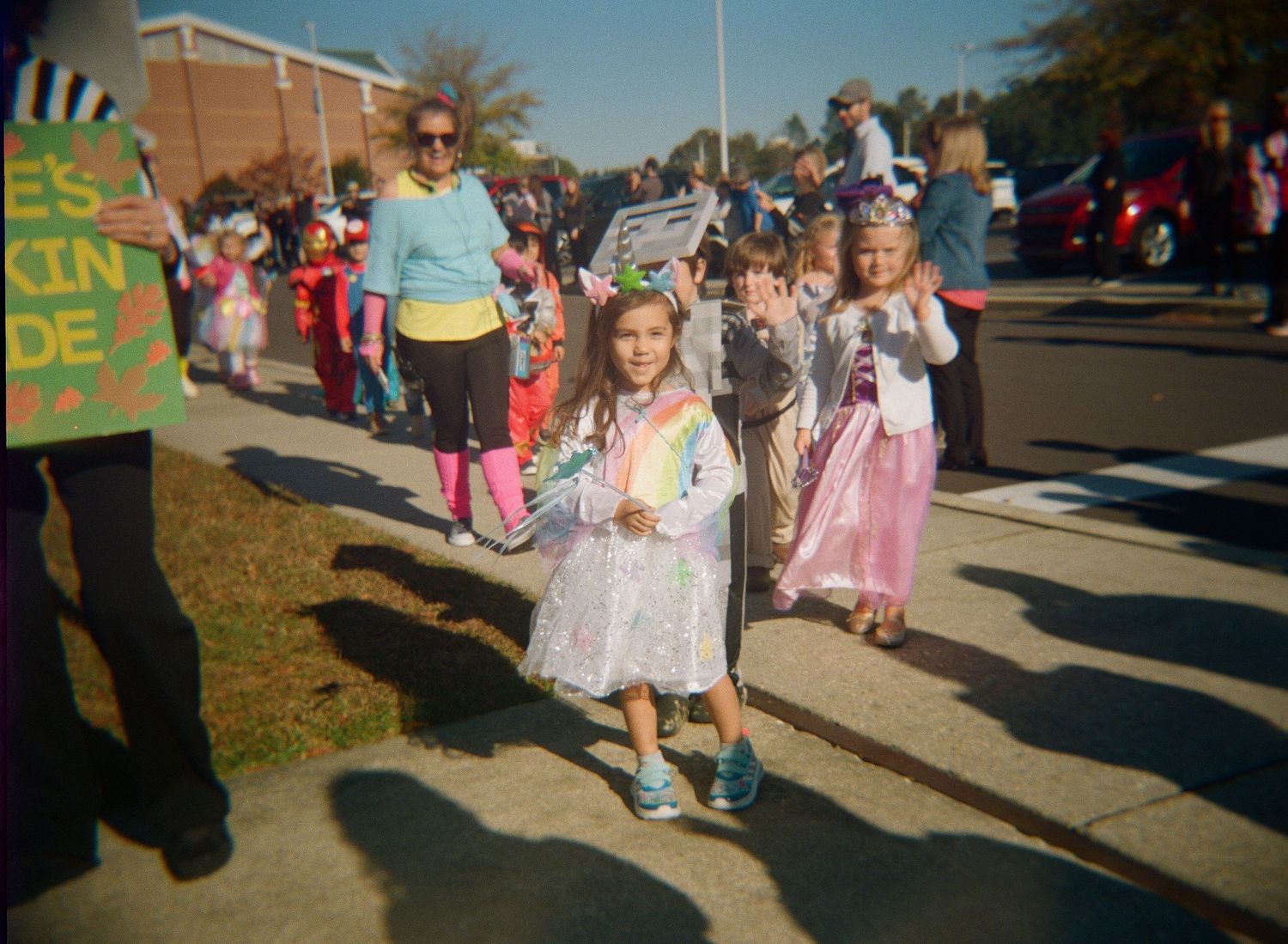 Children participate in a school parade wearing costumes while walking along a sidewalk on a sunny day.