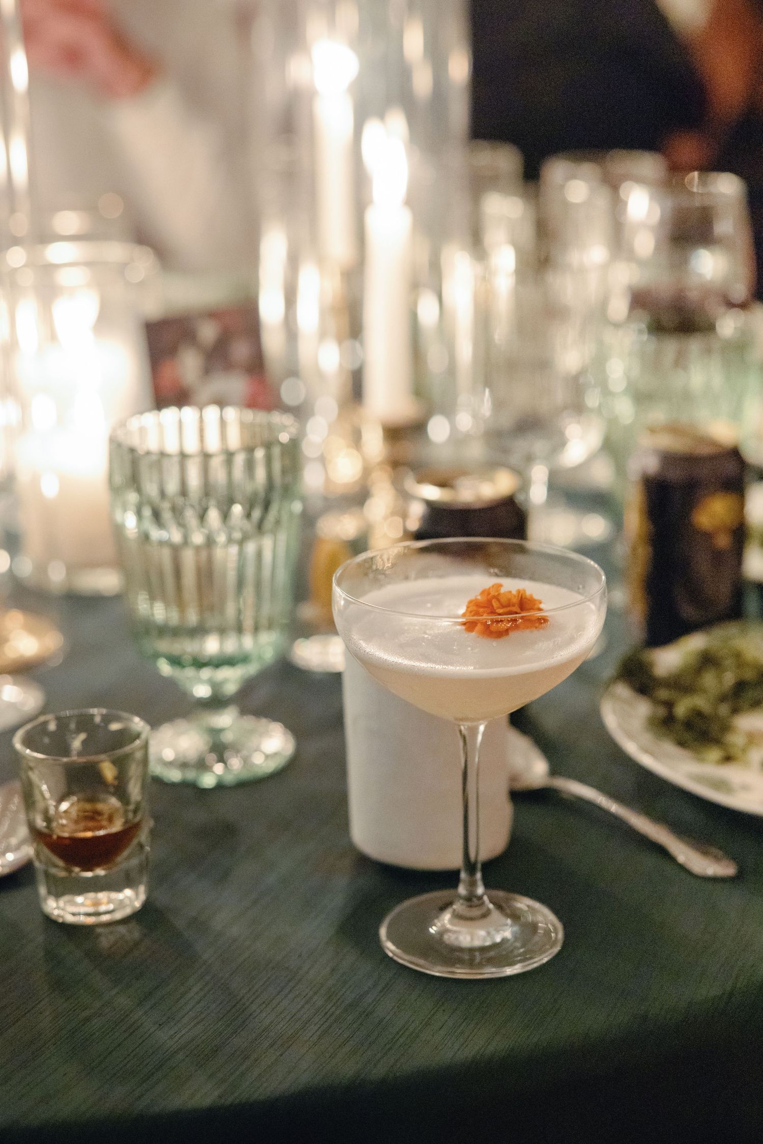 Elegant cocktail in coupe glass with orange garnish on candlelit dinner table with glassware.