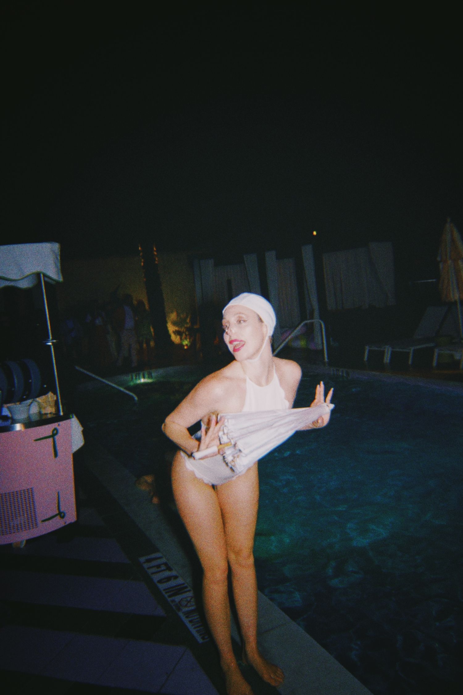 Woman in white swimsuit and swim cap poses playfully by illuminated pool at night.