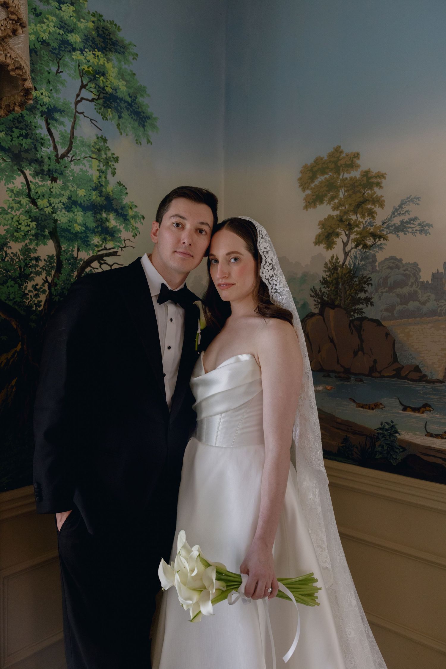 A couple in formal wedding attire poses for portraits in front of a painted scenic backdrop at dusk.