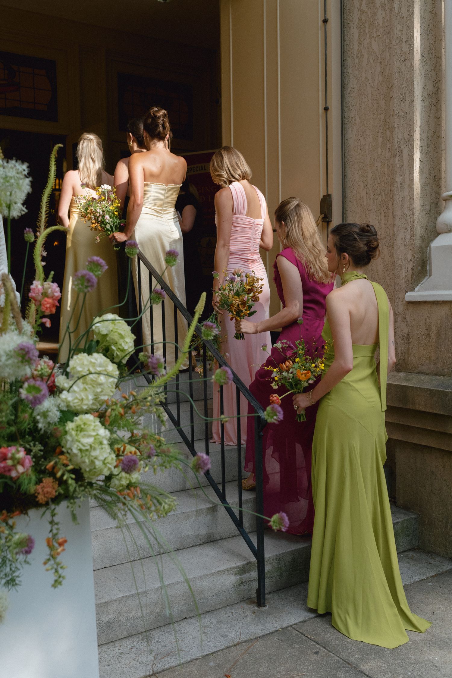 Bridesmaids in colorful gowns holding bouquets ascending stone steps into a venue for a wedding ceremony.