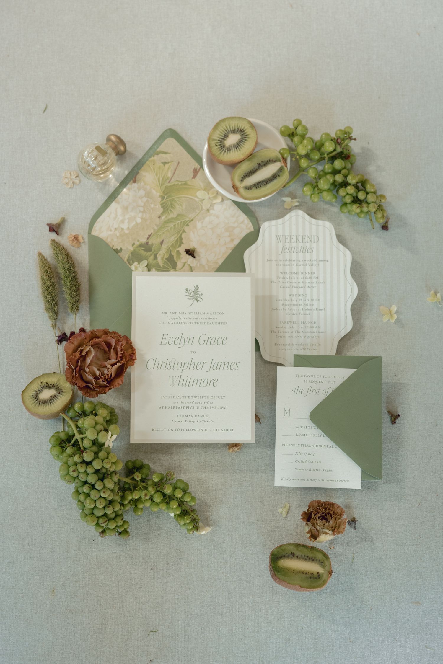 Elegant sage green wedding invitation suite with kiwi fruit, grapes, and dried florals on a light blue background.