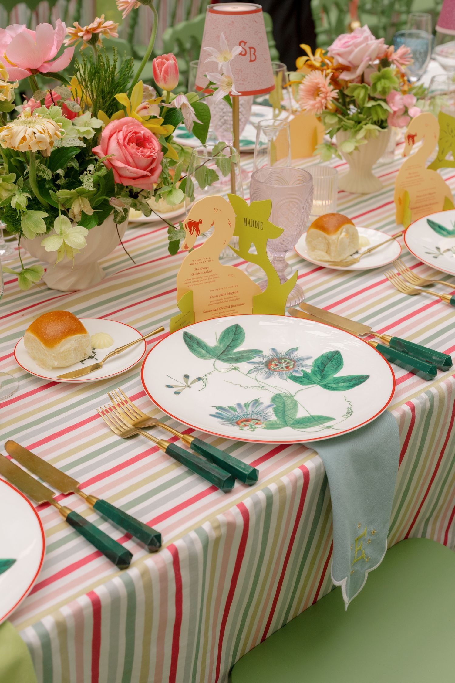 Elegant garden party table setting with floral plates, green-handled gold cutlery, striped tablecloth, and pink rose centerpieces.