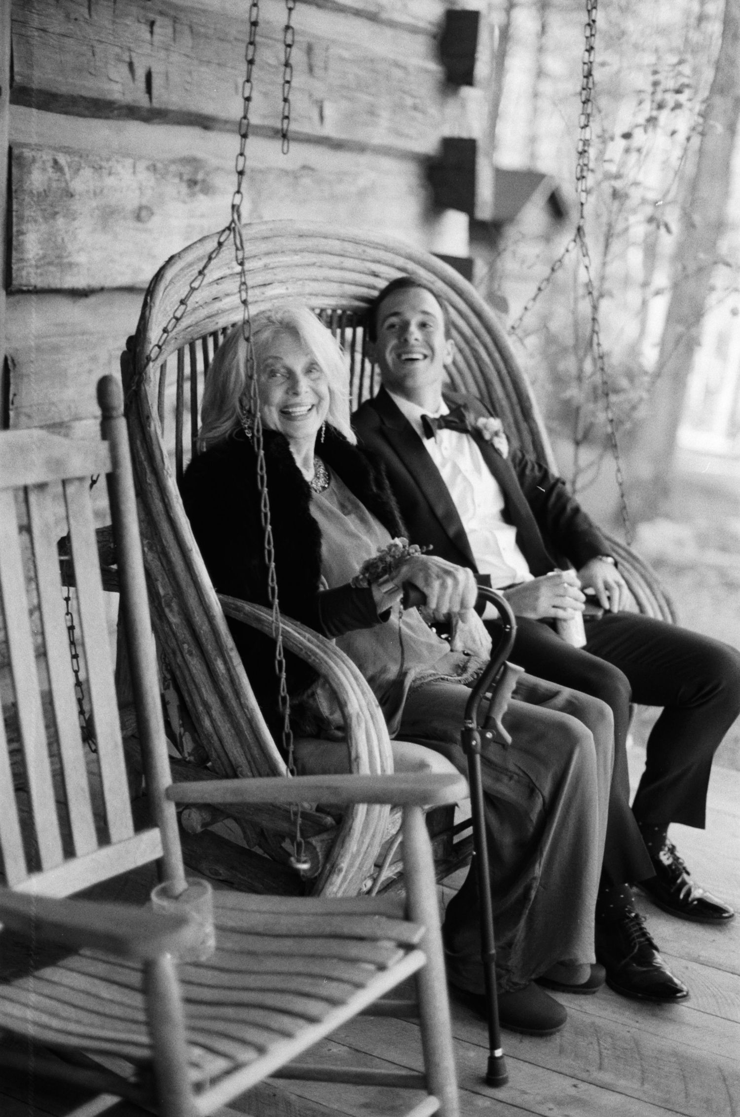 Elderly woman and man in tuxedo smiling on rustic log cabin porch swing in black and white photo.