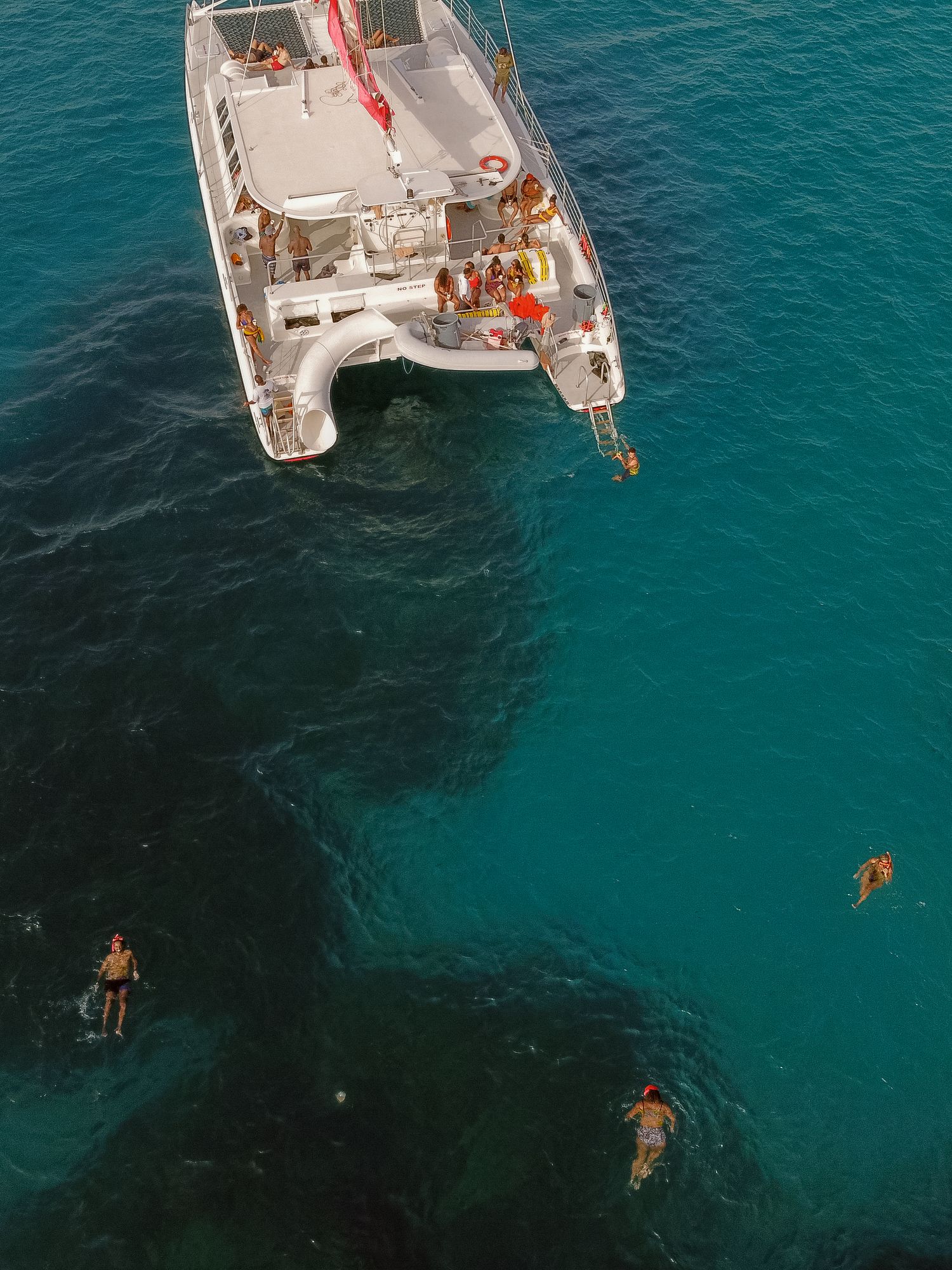 Aerial view of a white catamaran anchored in turquoise waters with swimmers snorkeling around the boat.