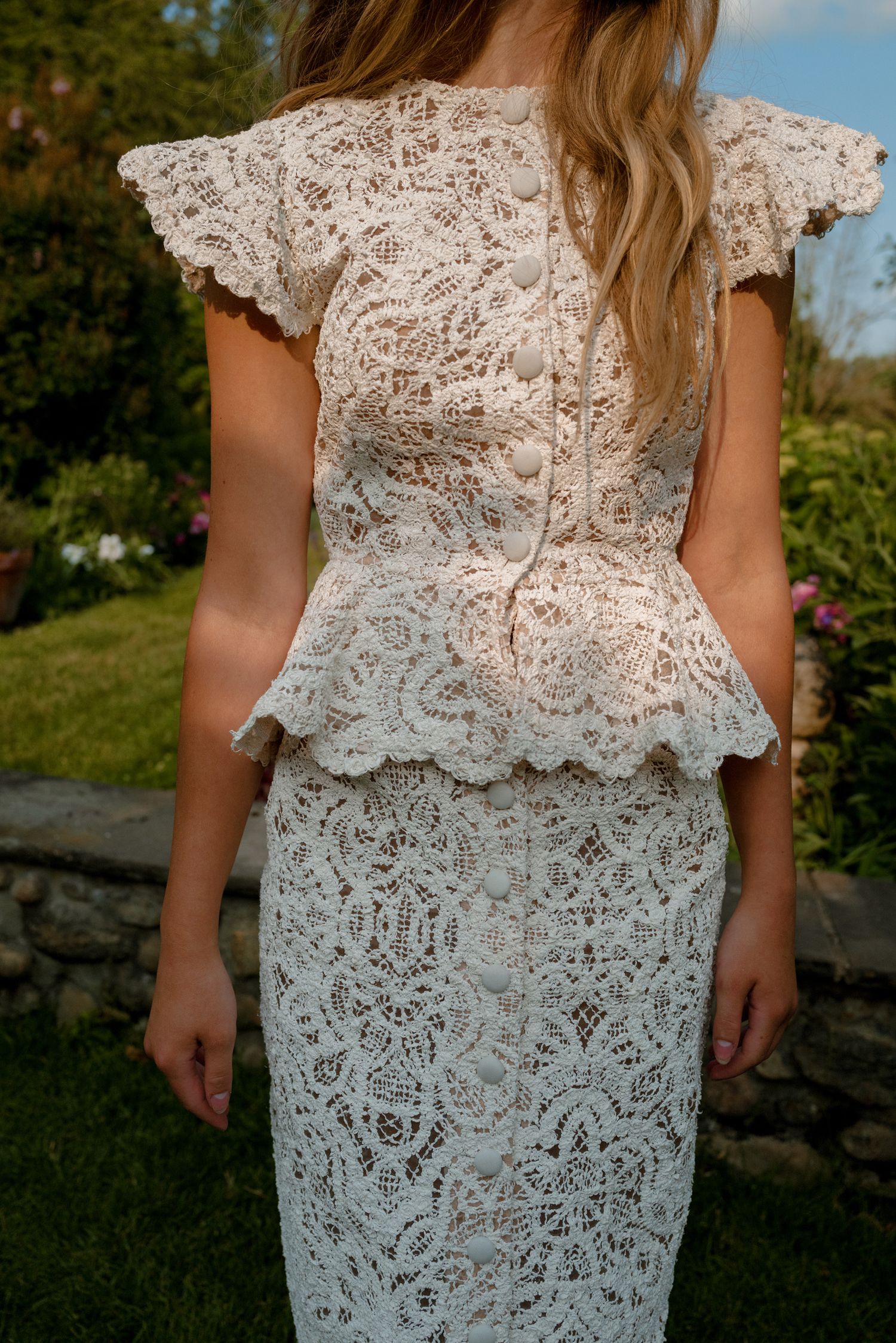 Back view of woman wearing elegant white lace peplum top and matching midi skirt in garden.