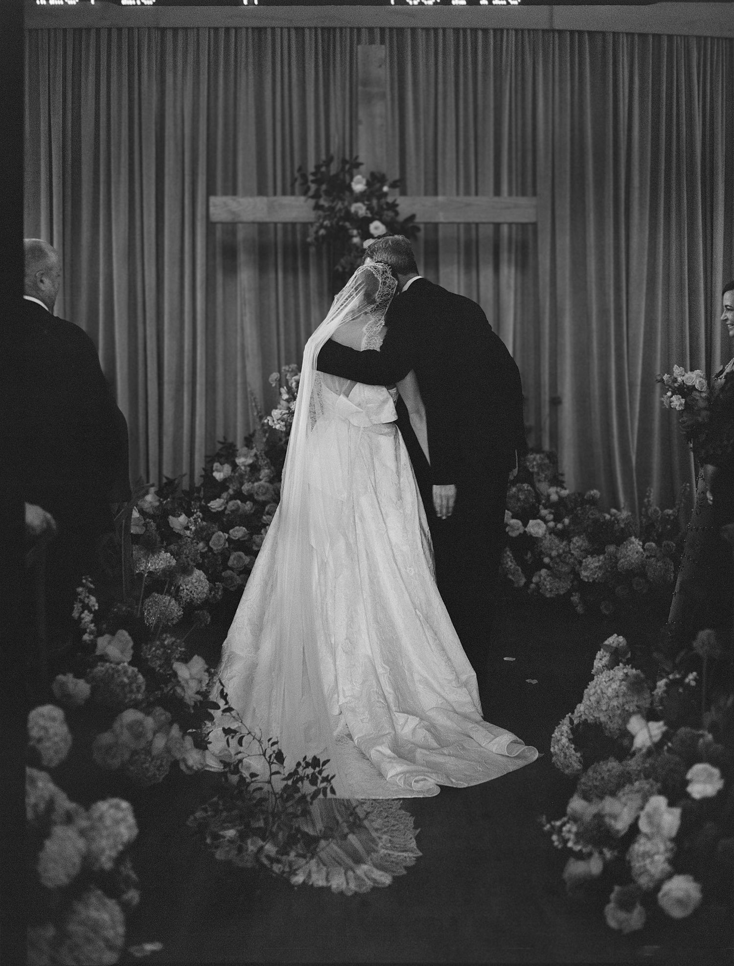 Black and white photo of a bride and groom kissing at the altar, surrounded by floral arrangements and a cross.