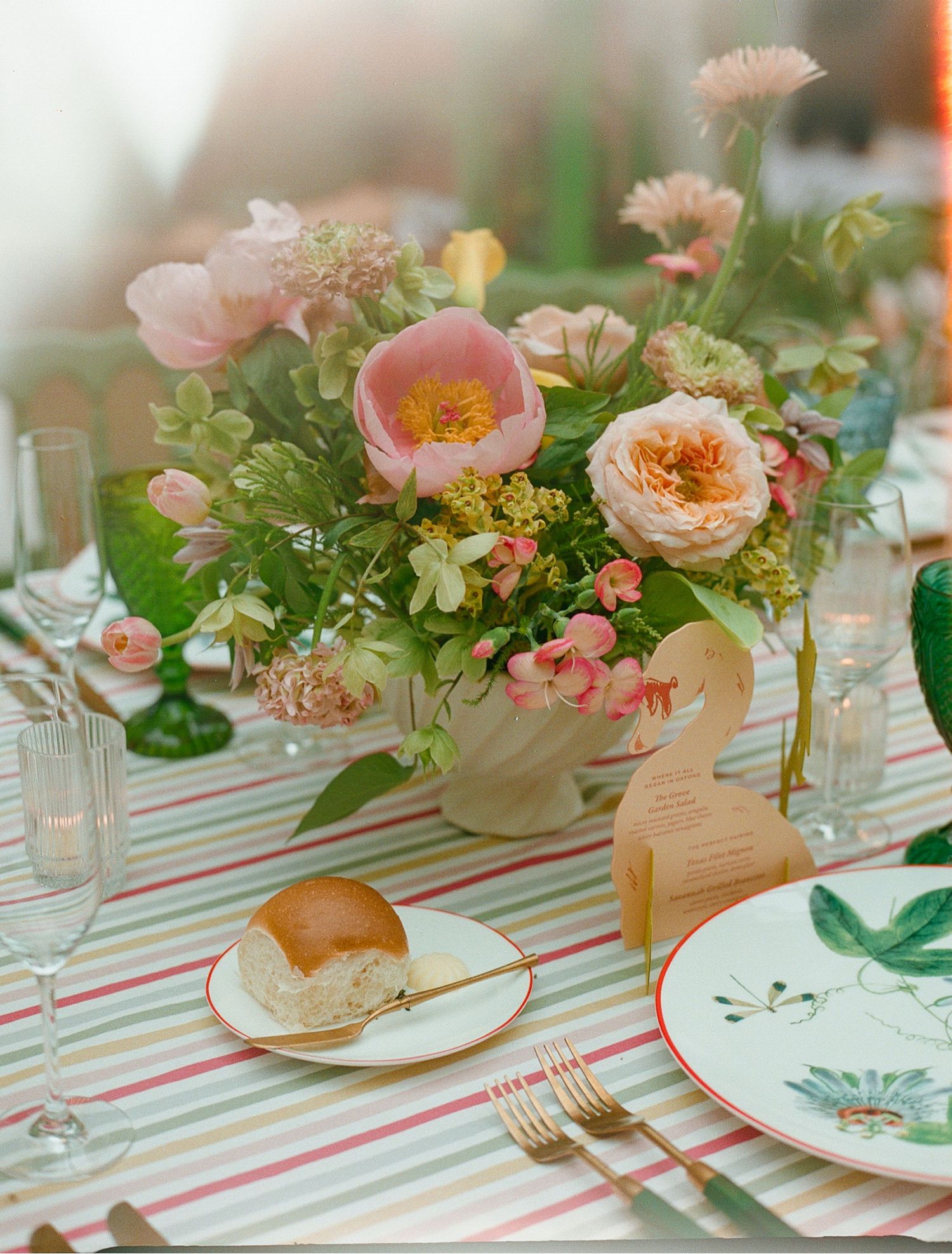 Elegant wedding tablescape with pink peony floral centerpiece, striped linen, gold flatware, and white plates with a dinner roll.
