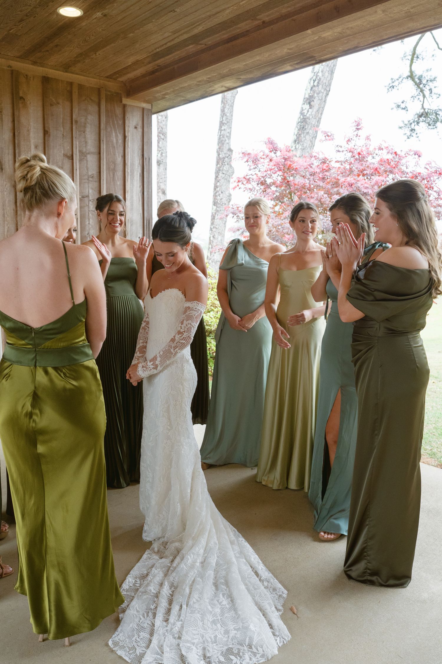 Bride in lace gown shares emotional reveal moment with bridesmaids in sage and olive green satin dresses.