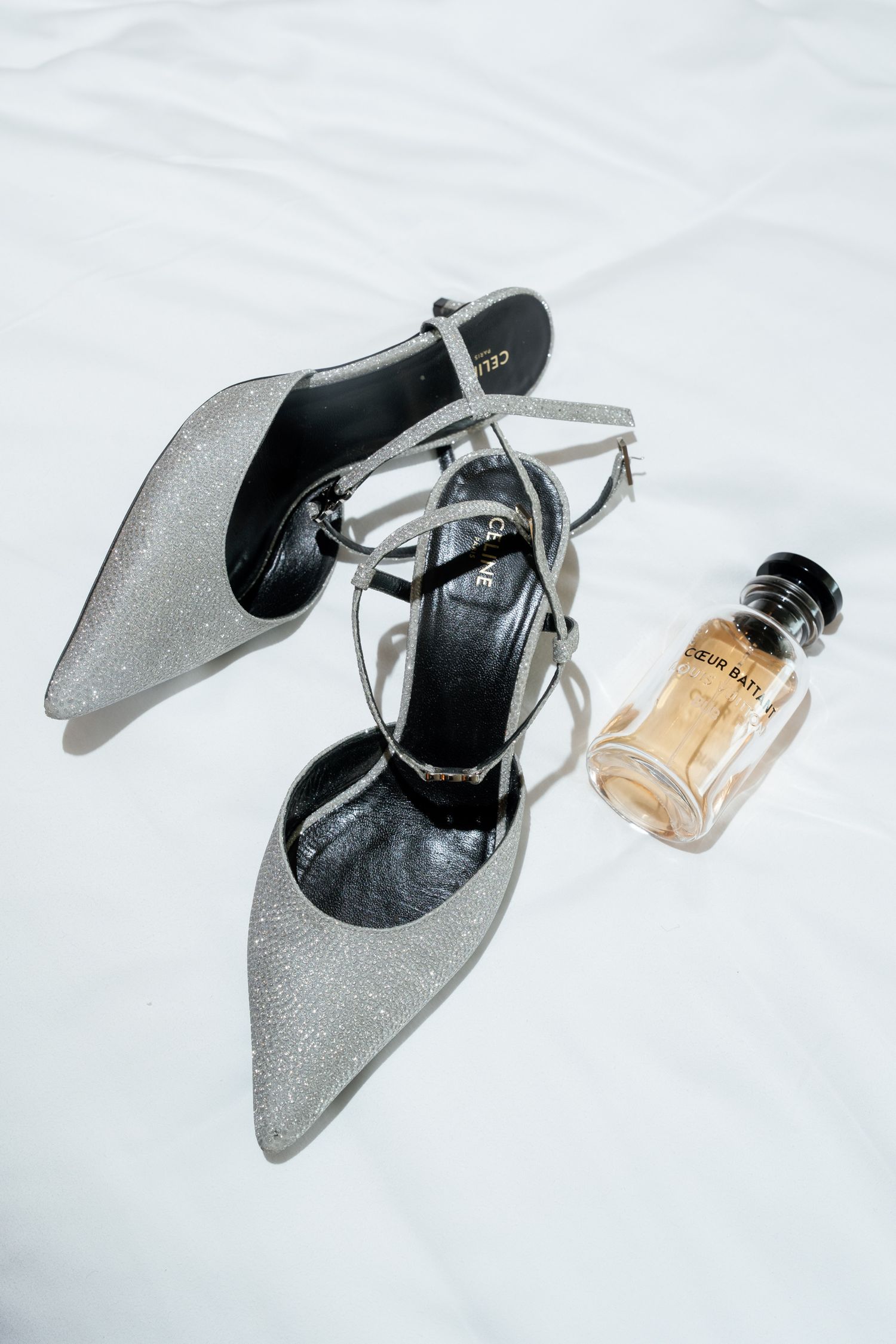 Silver glitter stiletto heels with ankle straps beside a small perfume bottle on white fabric.