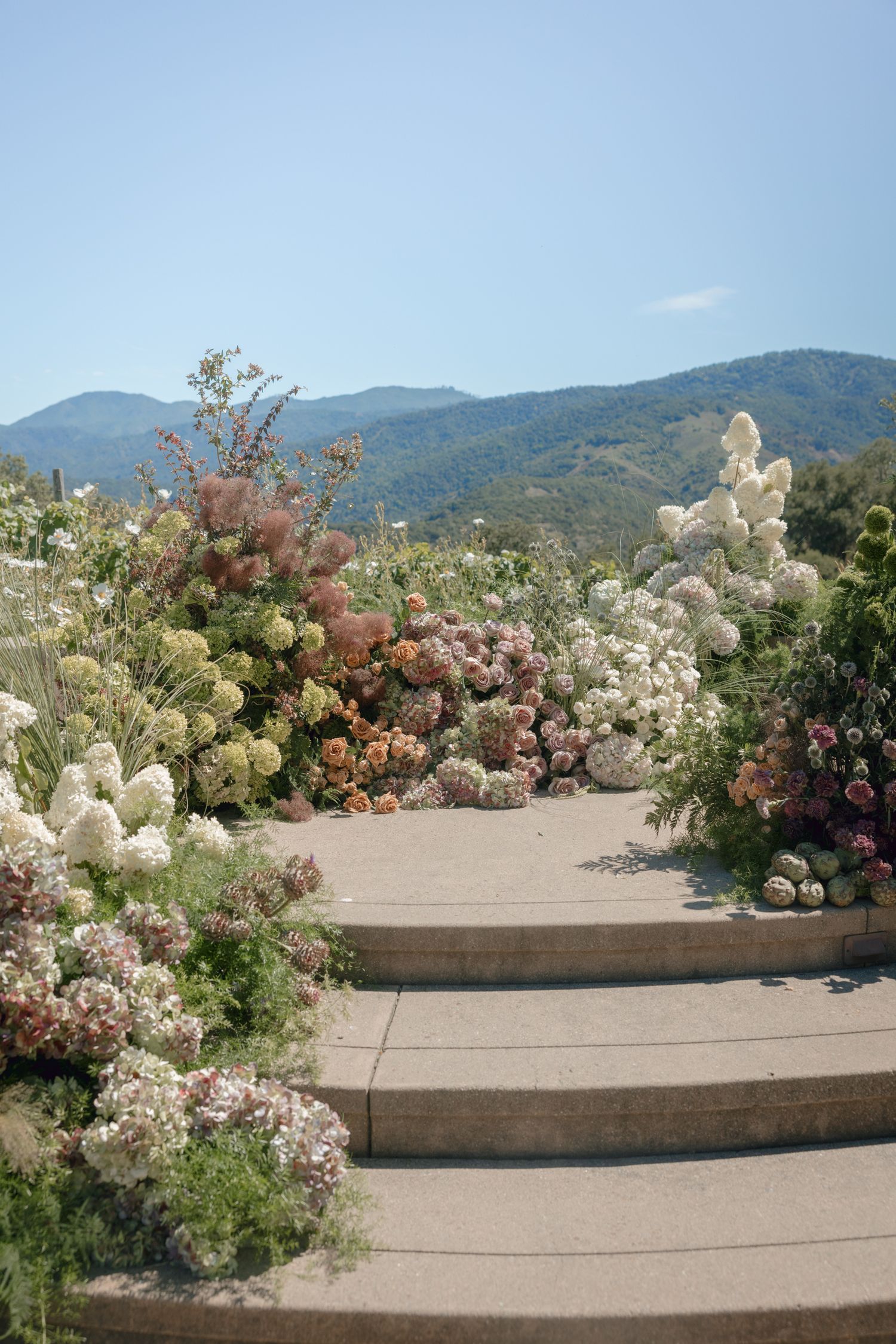 Terraced concrete steps adorned with pink and white flowering bushes against a mountain backdrop under blue sky.