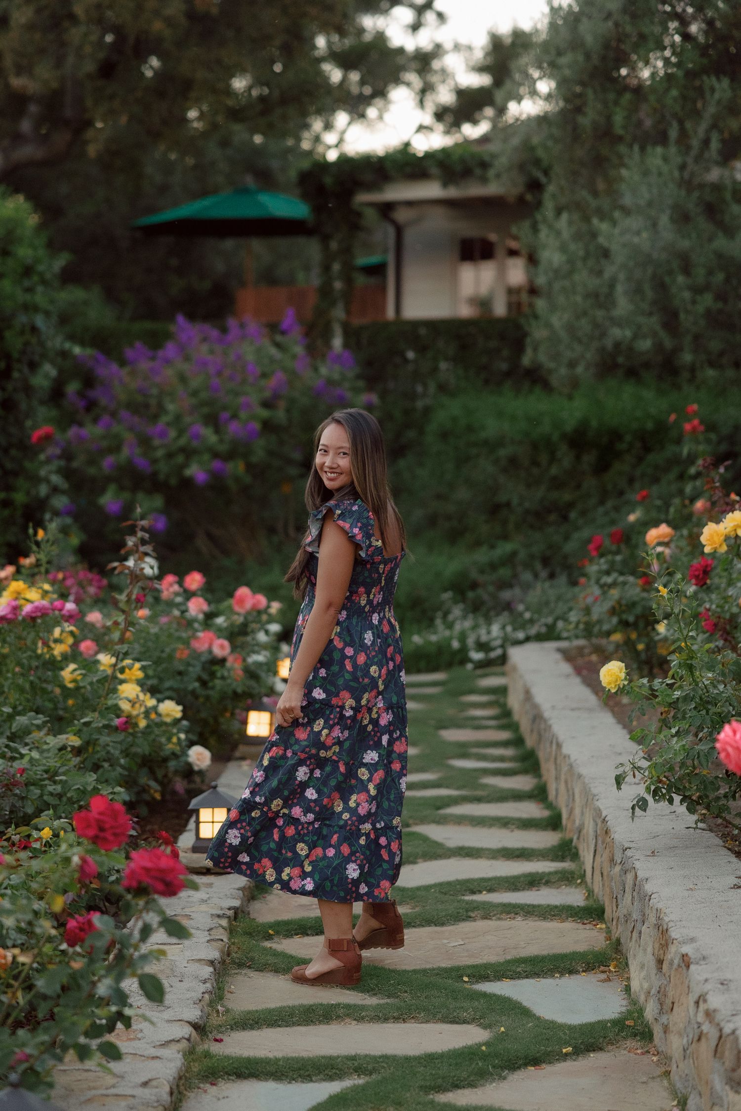 A stone pathway winds through a colorful rose garden with blooming flowers in pink, yellow and purple hues.