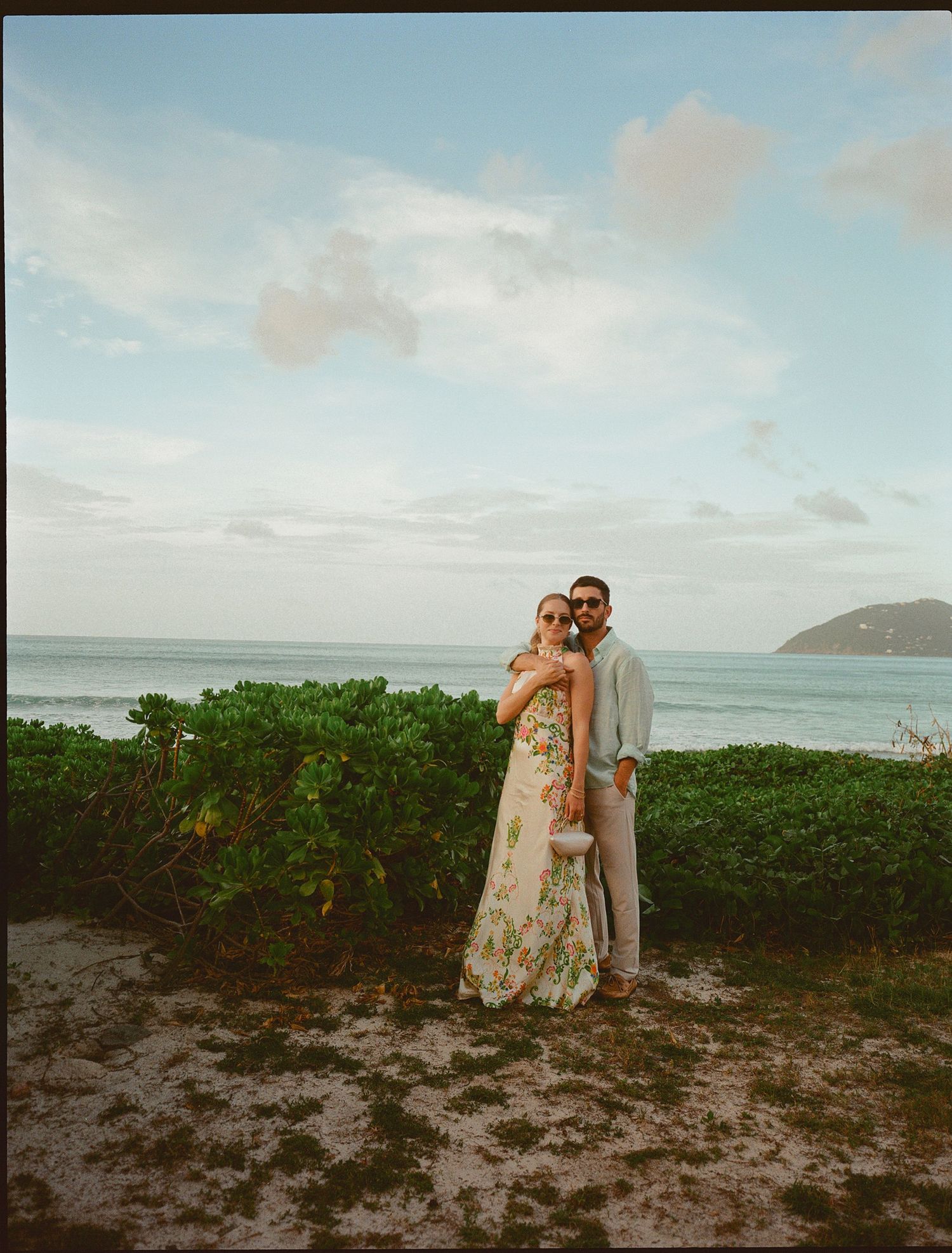 Couple posing together near lush green hedges with ocean and blue sky backdrop at a tropical coastal location.