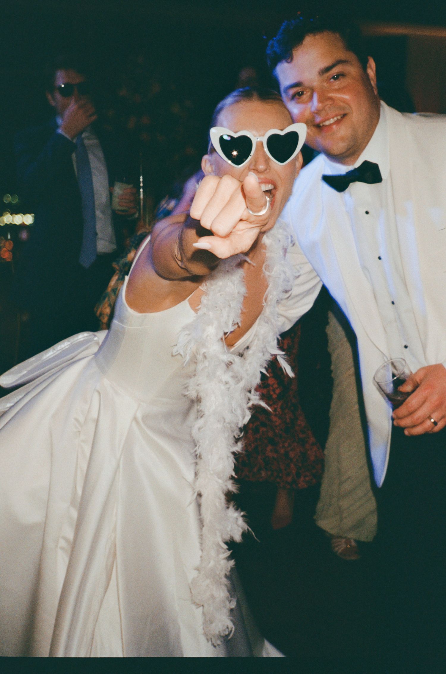 A couple in wedding attire dances at their reception, with the bride wearing heart-shaped sunglasses and a feather boa.