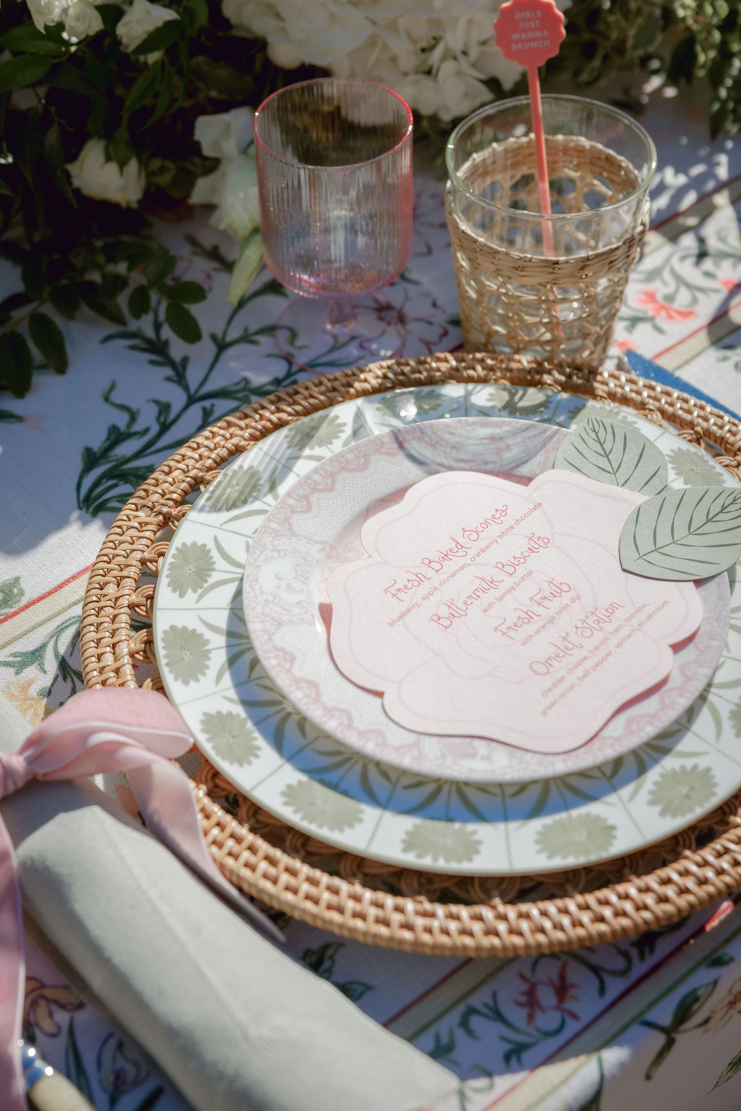 Place settings with floral patterned plates, woven copper chargers, and pink accents on a blue tablecloth.
